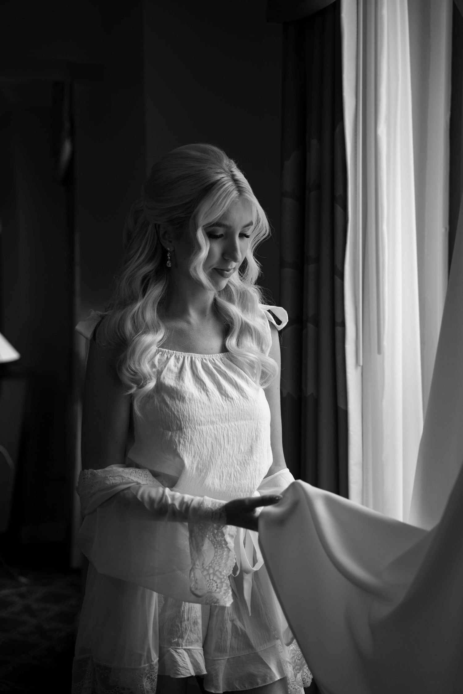 Black and white portrait of a smiling blonde bride in a white top holding her wedding dress by a window.