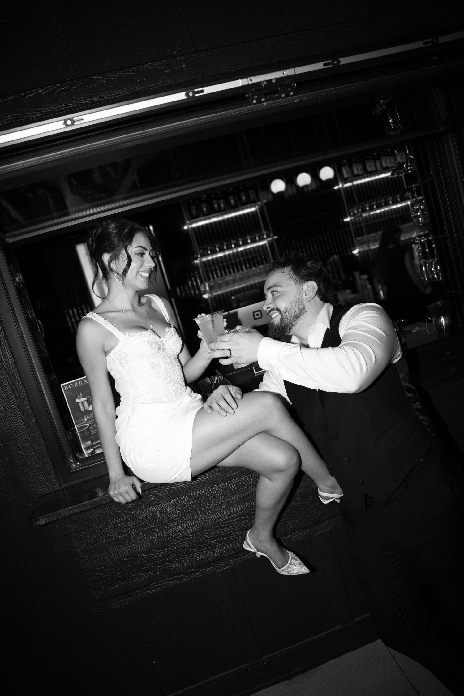 Black and white photo of a couple in wedding attire toasting drinks at a bar, sharing a joyful moment.