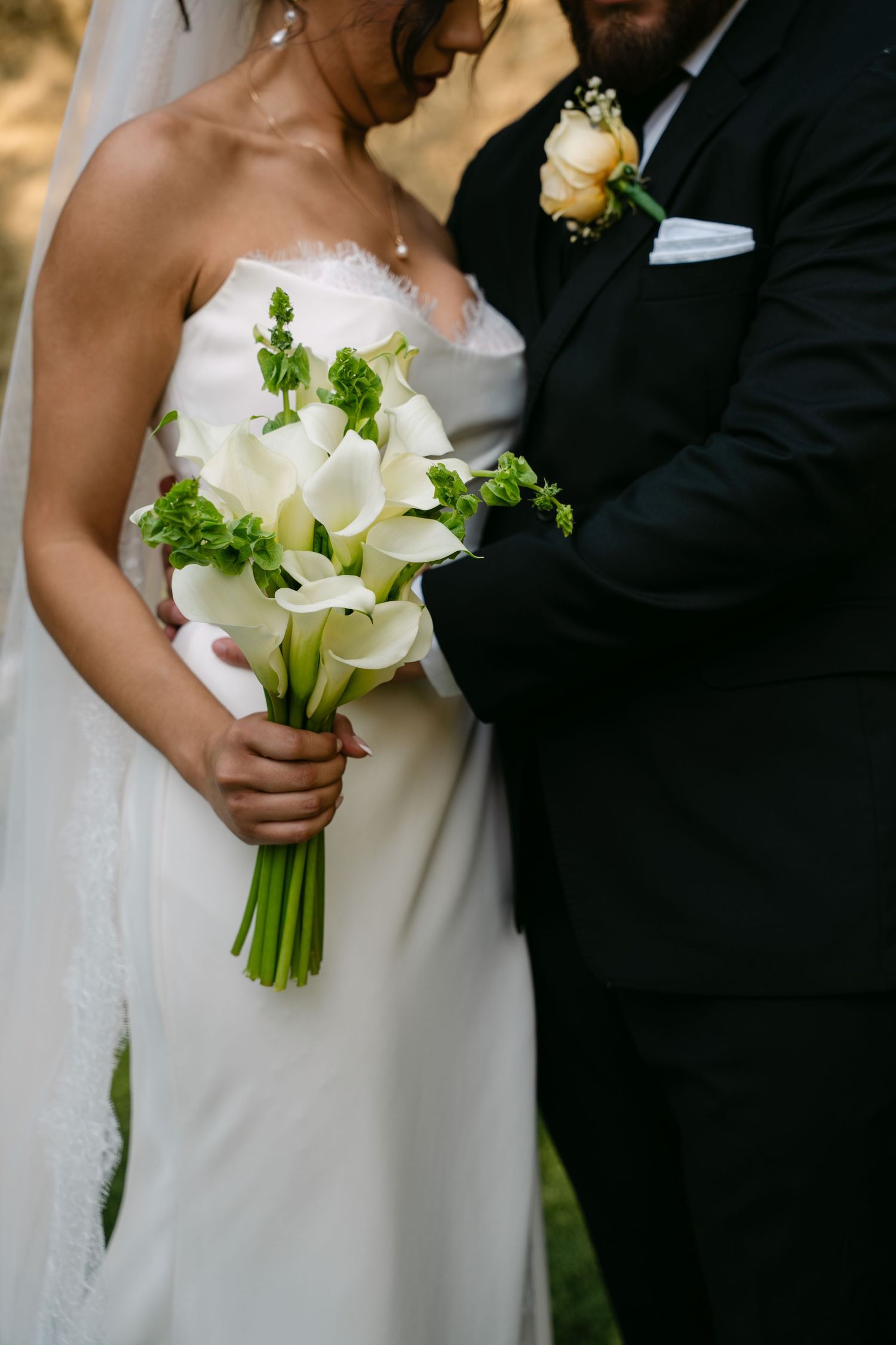 Bride holds elegant white calla lily bouquet with green accents beside groom in black suit.
