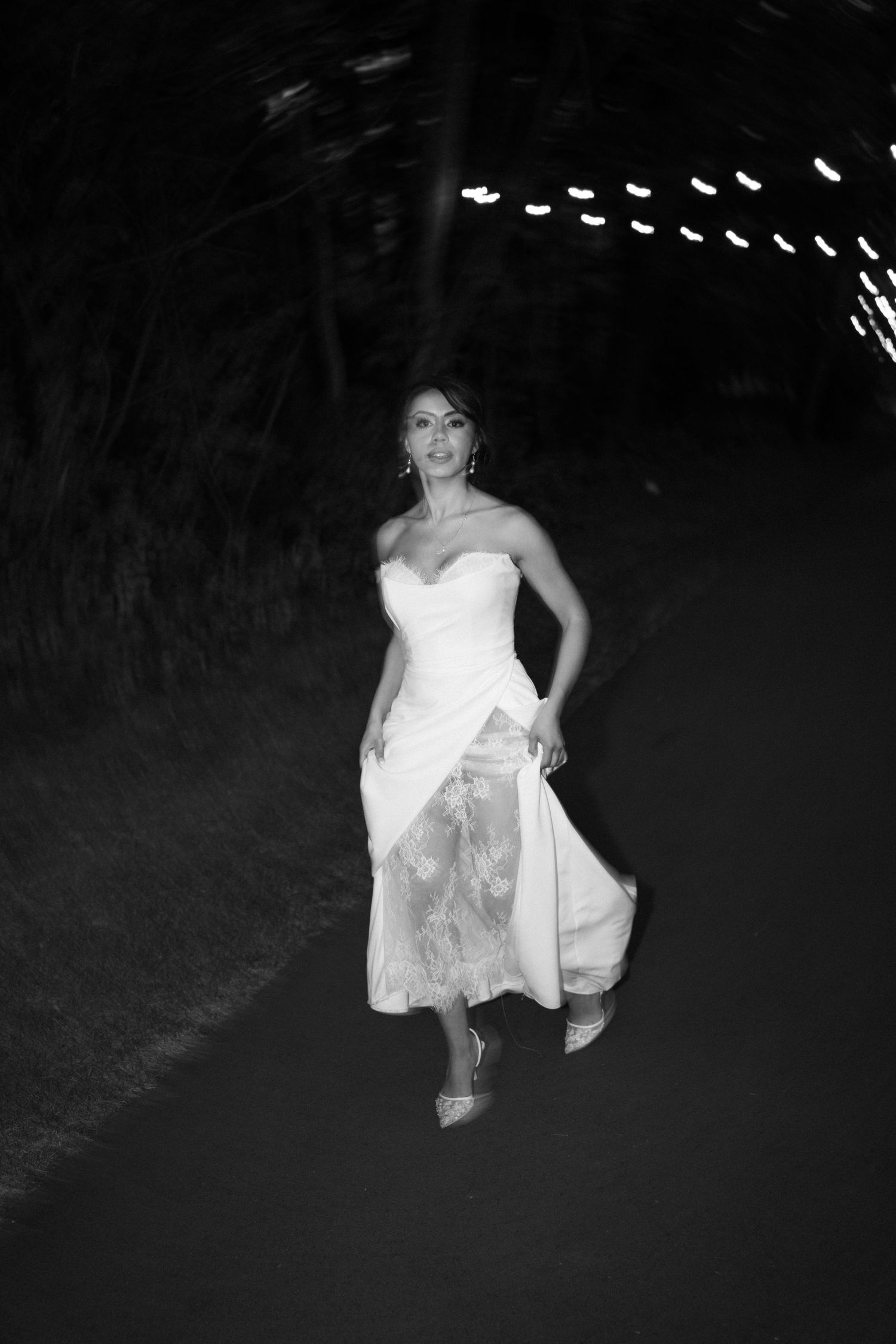 Black and white image of bride in strapless white lace gown walking confidently on dark pathway.