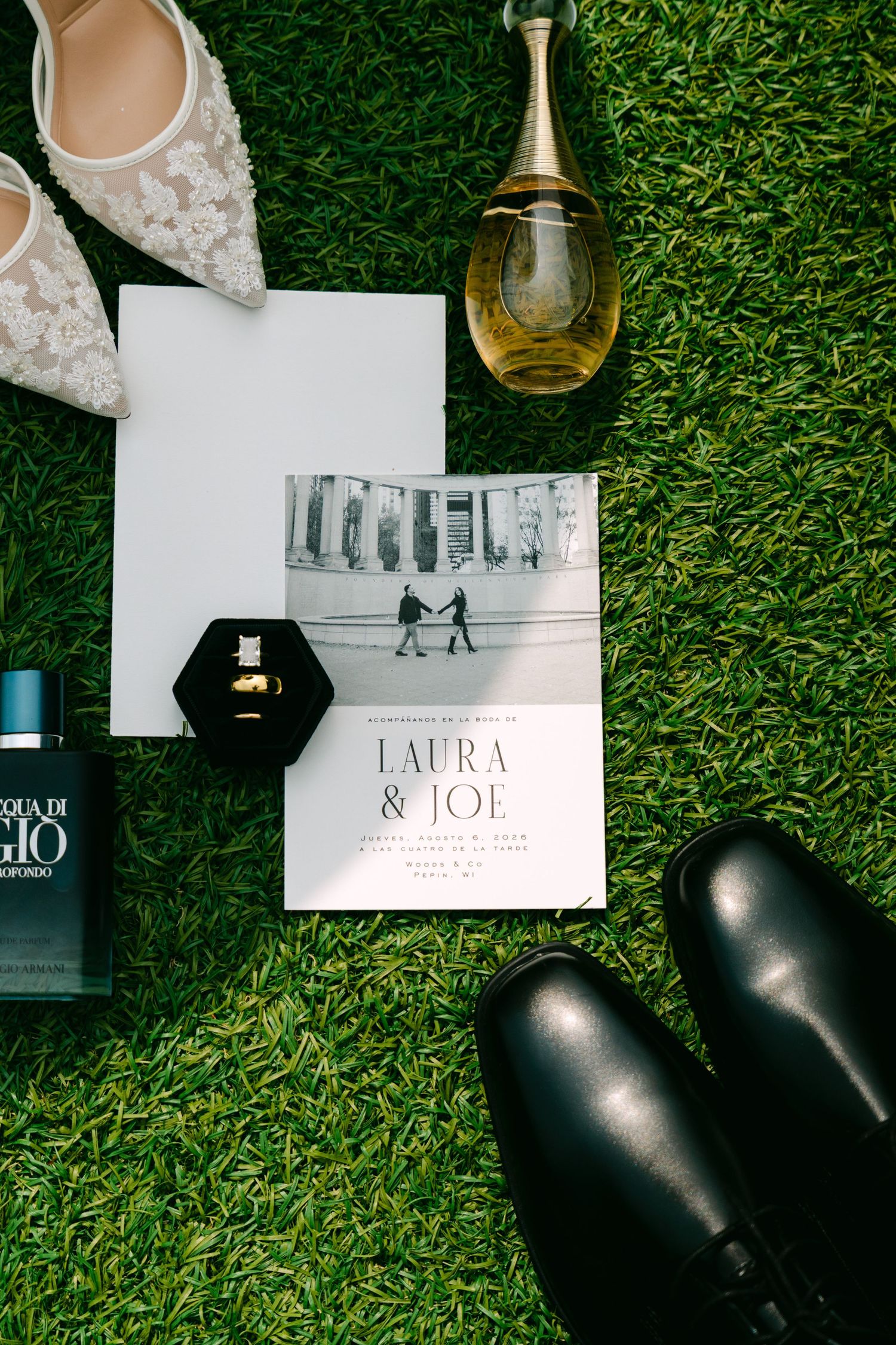 Overhead flat lay of wedding details including invitation, perfume, rings, bridal heels, and dress shoes on grass.
