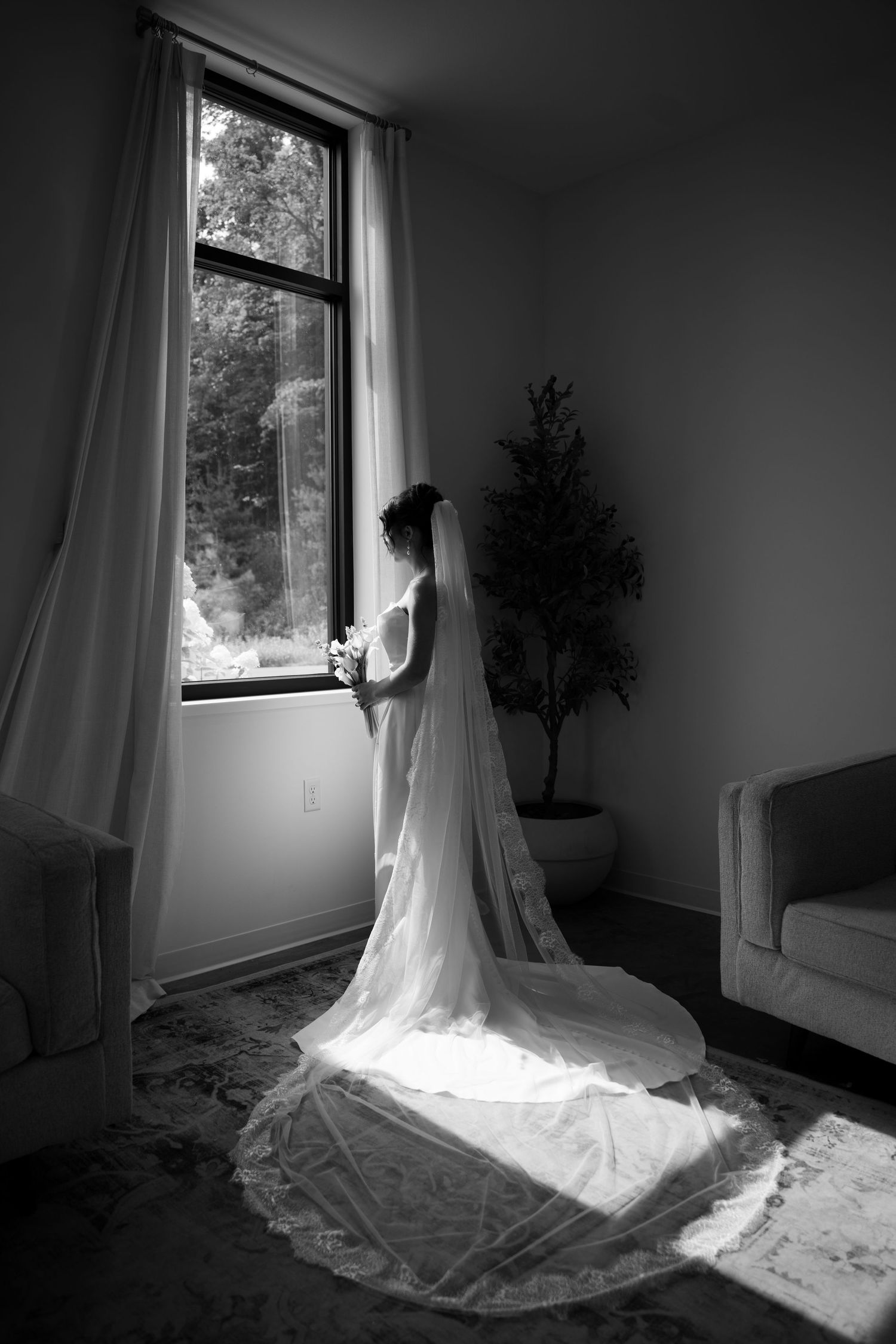 Black and white silhouette of a bride in a wedding gown with cathedral veil by a window.