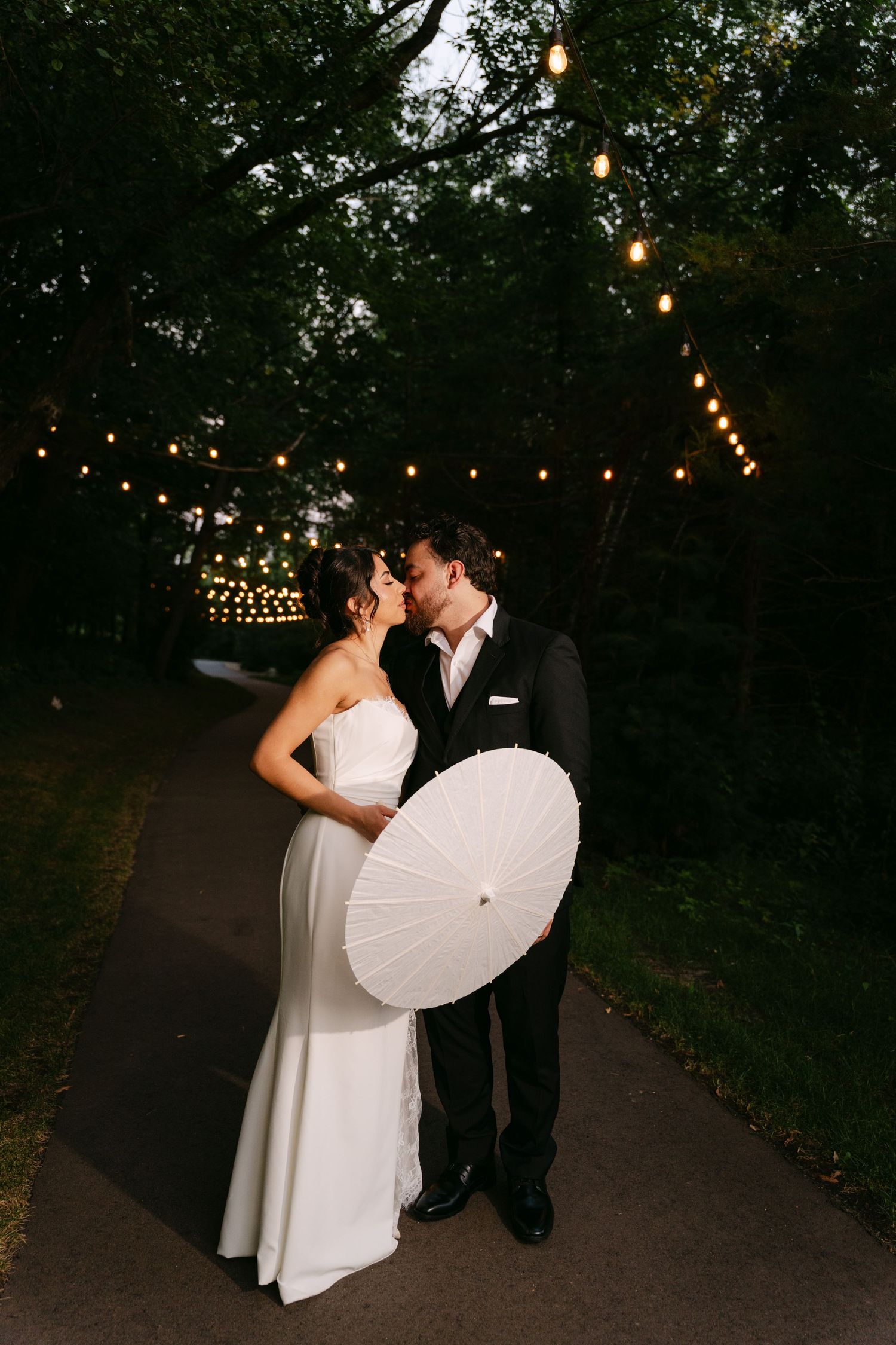 Wedding couple kissing under glowing string lights at dusk, bride holding white paper parasol.