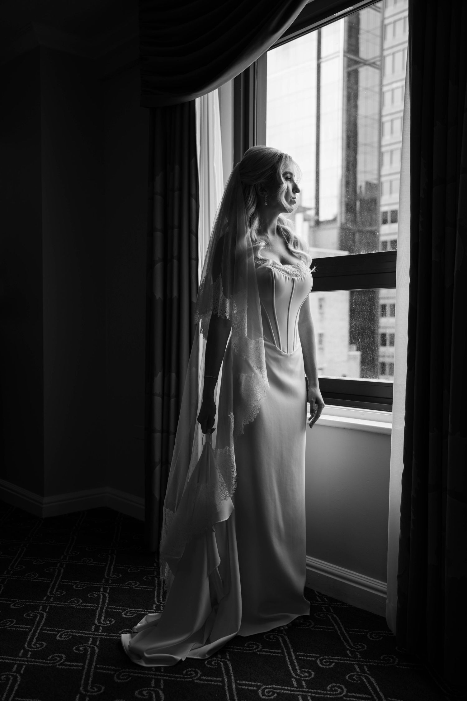 Elegant bride in white gown and veil stands by large window in black and white full-length portrait.