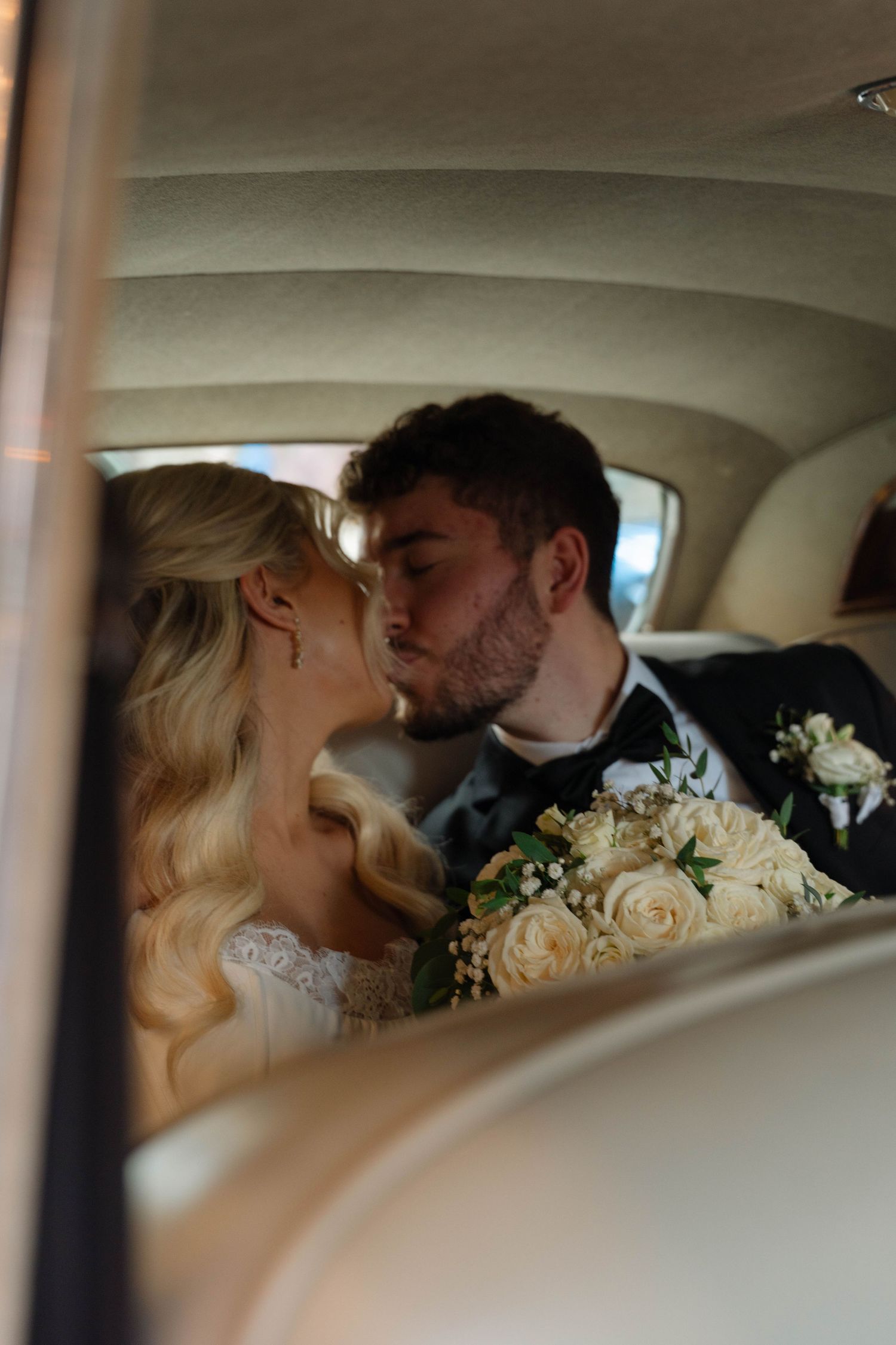 Bride and groom sharing a kiss inside a vintage car, bride holding white rose bouquet.