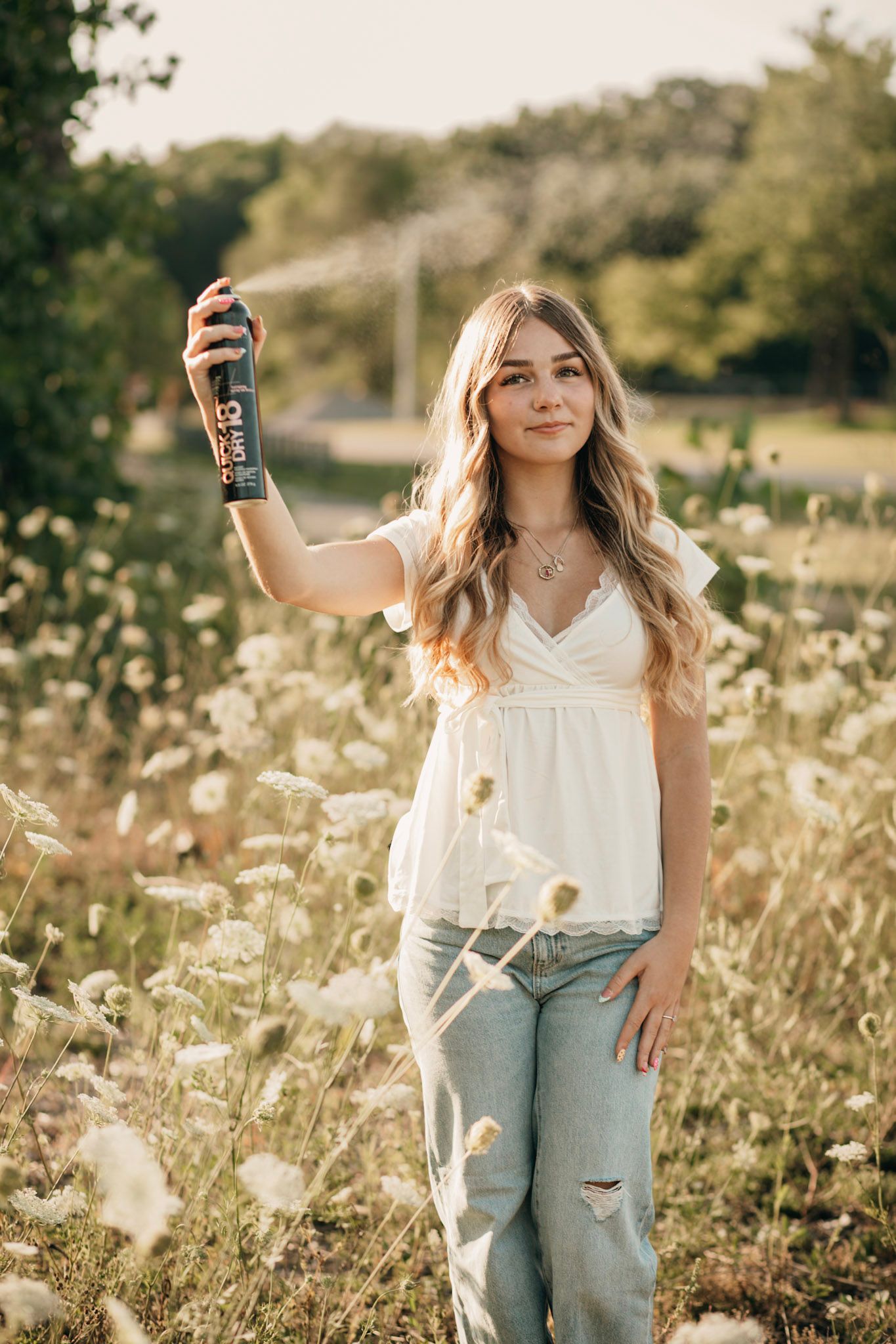 Young woman in white top and jeans holding hairspray in a sunlit wildflower field.