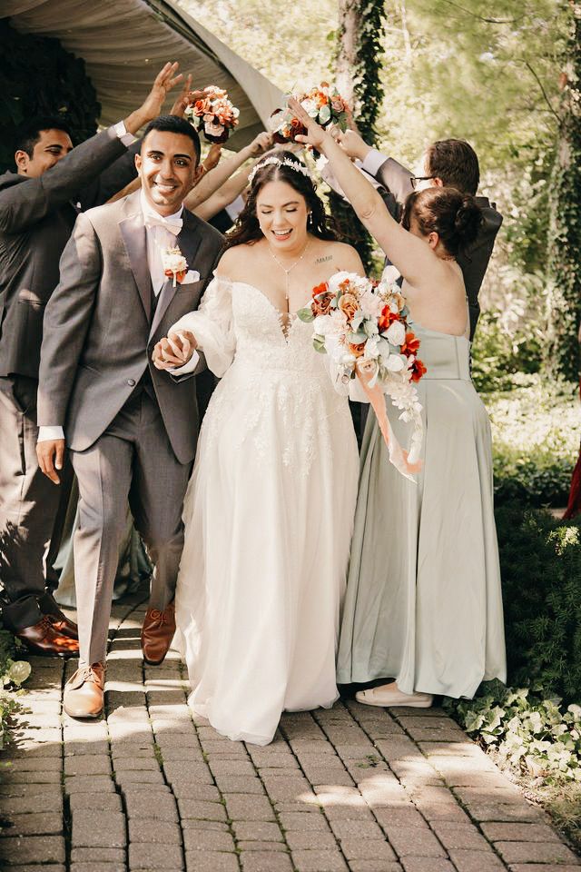 Bride and groom walk down aisle as wedding party celebrates with bouquets raised in outdoor garden ceremony.