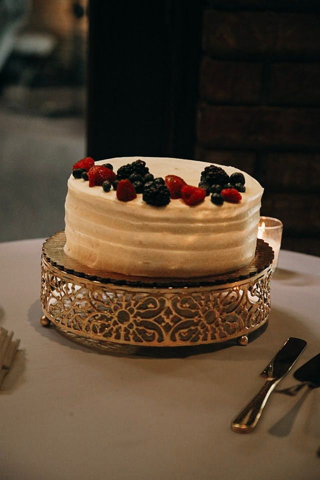 Single-tier white frosted wedding cake topped with fresh berries on ornate gold cake stand.