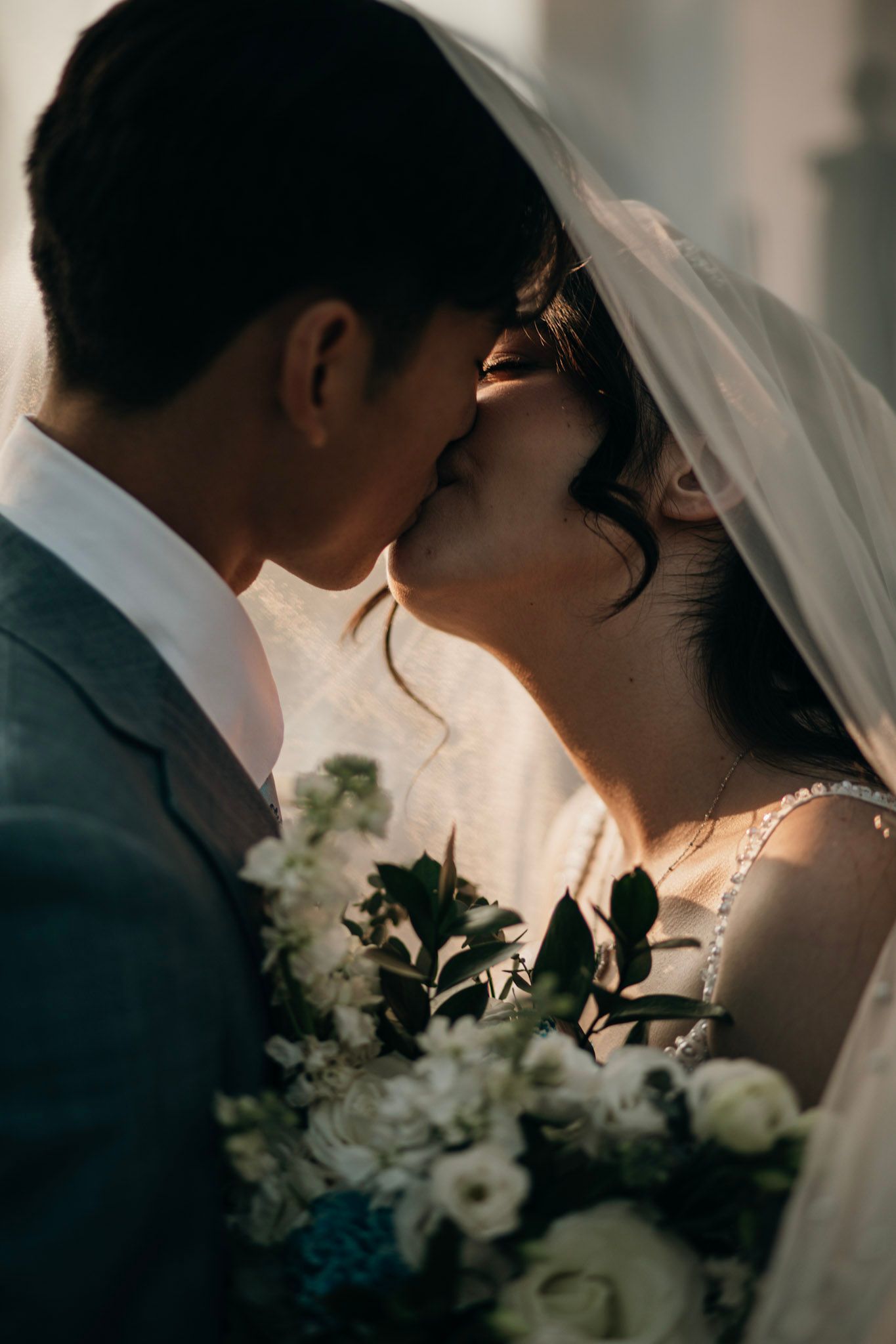 A bride and groom share a tender kiss under a white veil, with the bride holding a bouquet of white flowers.