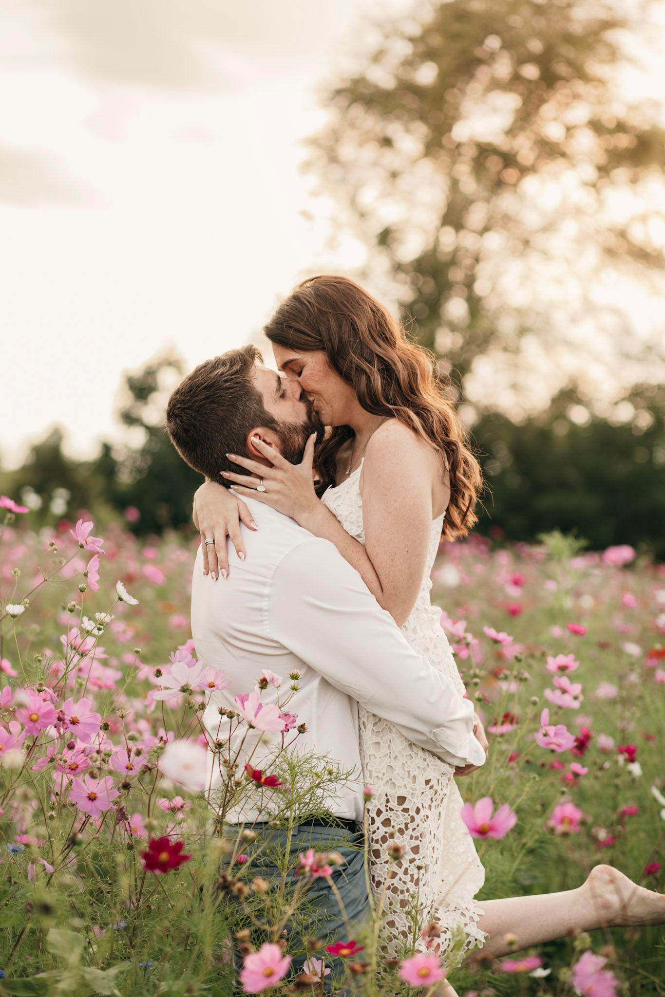 A couple shares a romantic kiss surrounded by blooming pink cosmos flowers in a sunlit field.