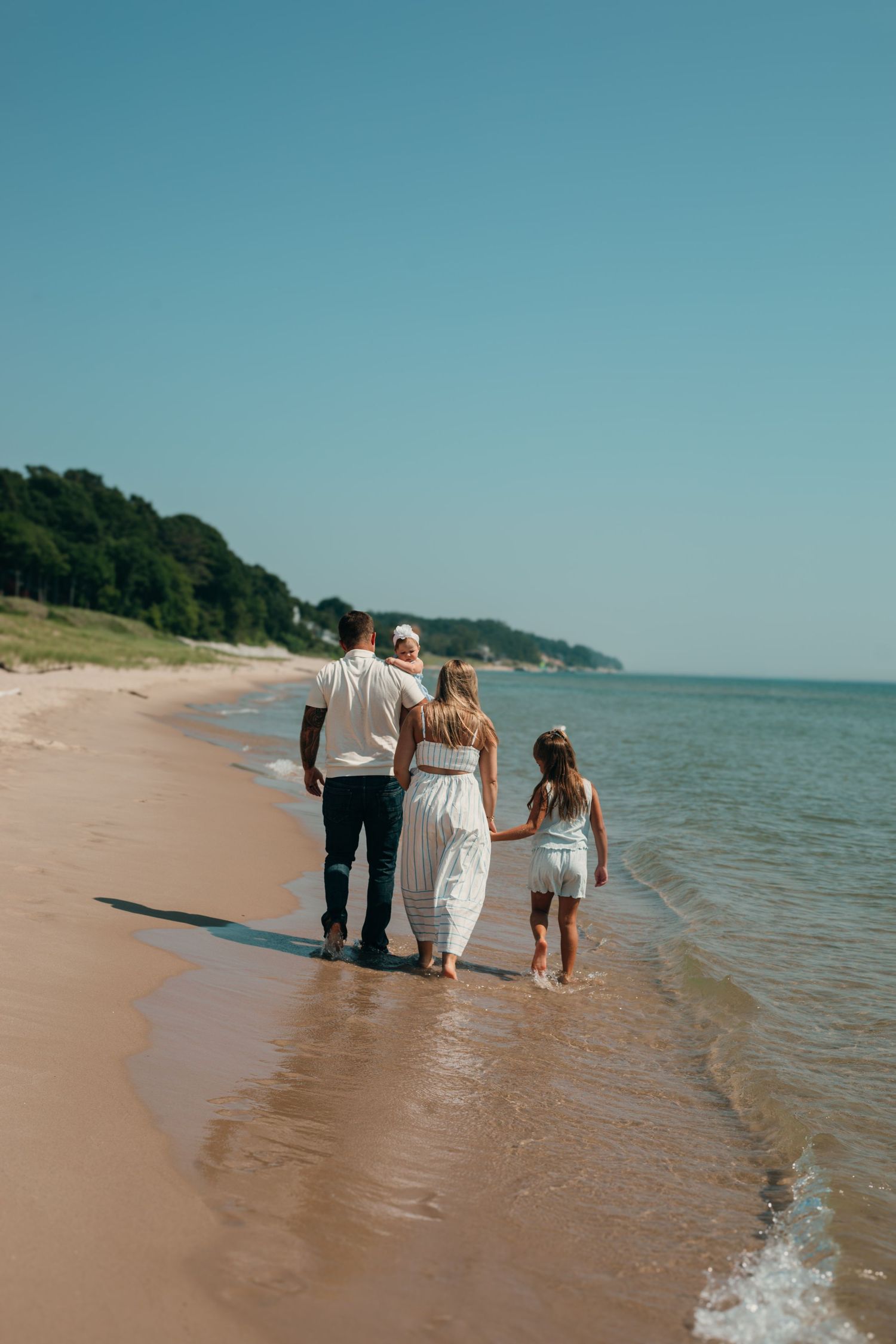 A family of three walks hand in hand along a sunny sandy beach shoreline.