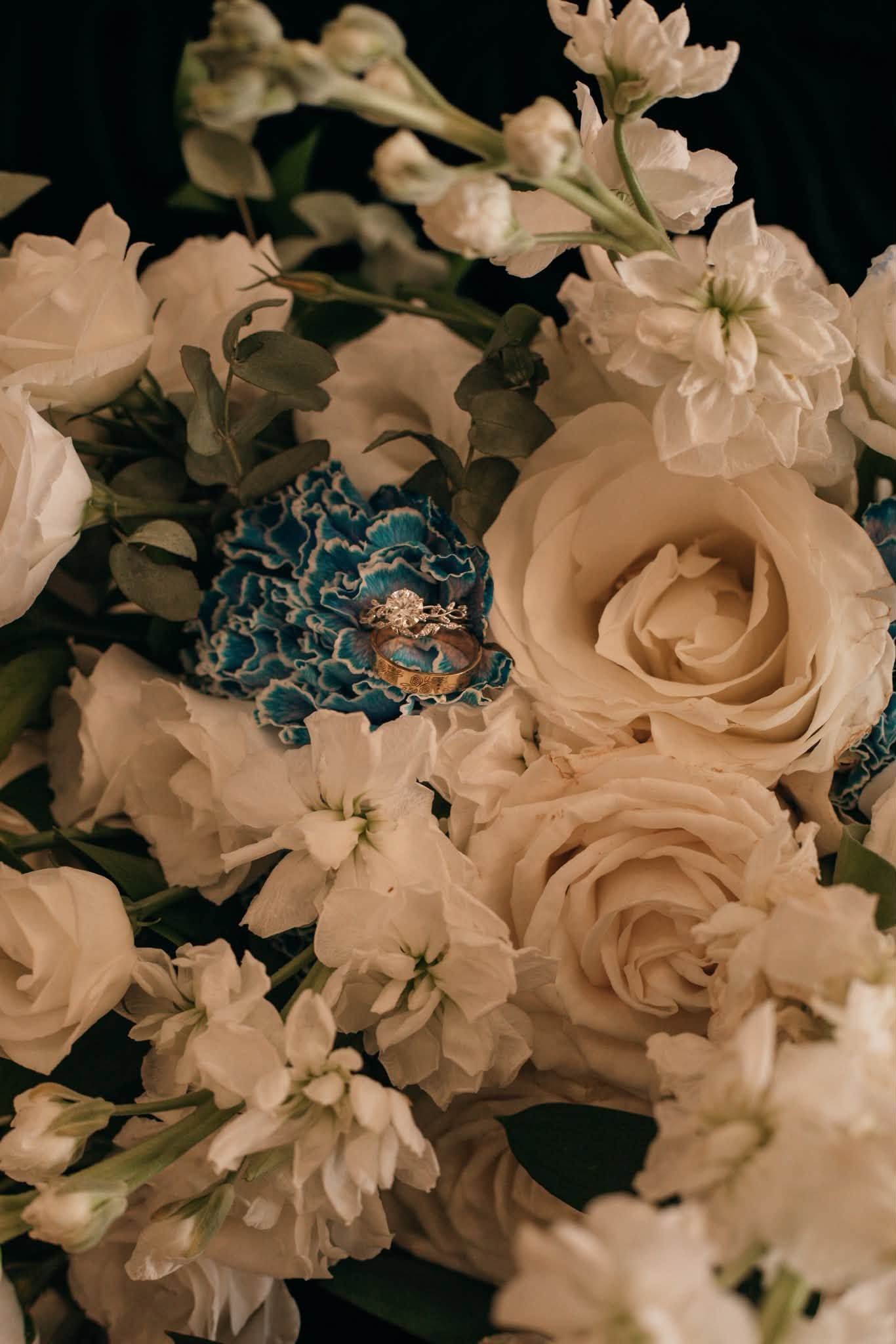 Gold engagement ring with diamond resting on blue lace among white roses and flowers in a bridal bouquet.
