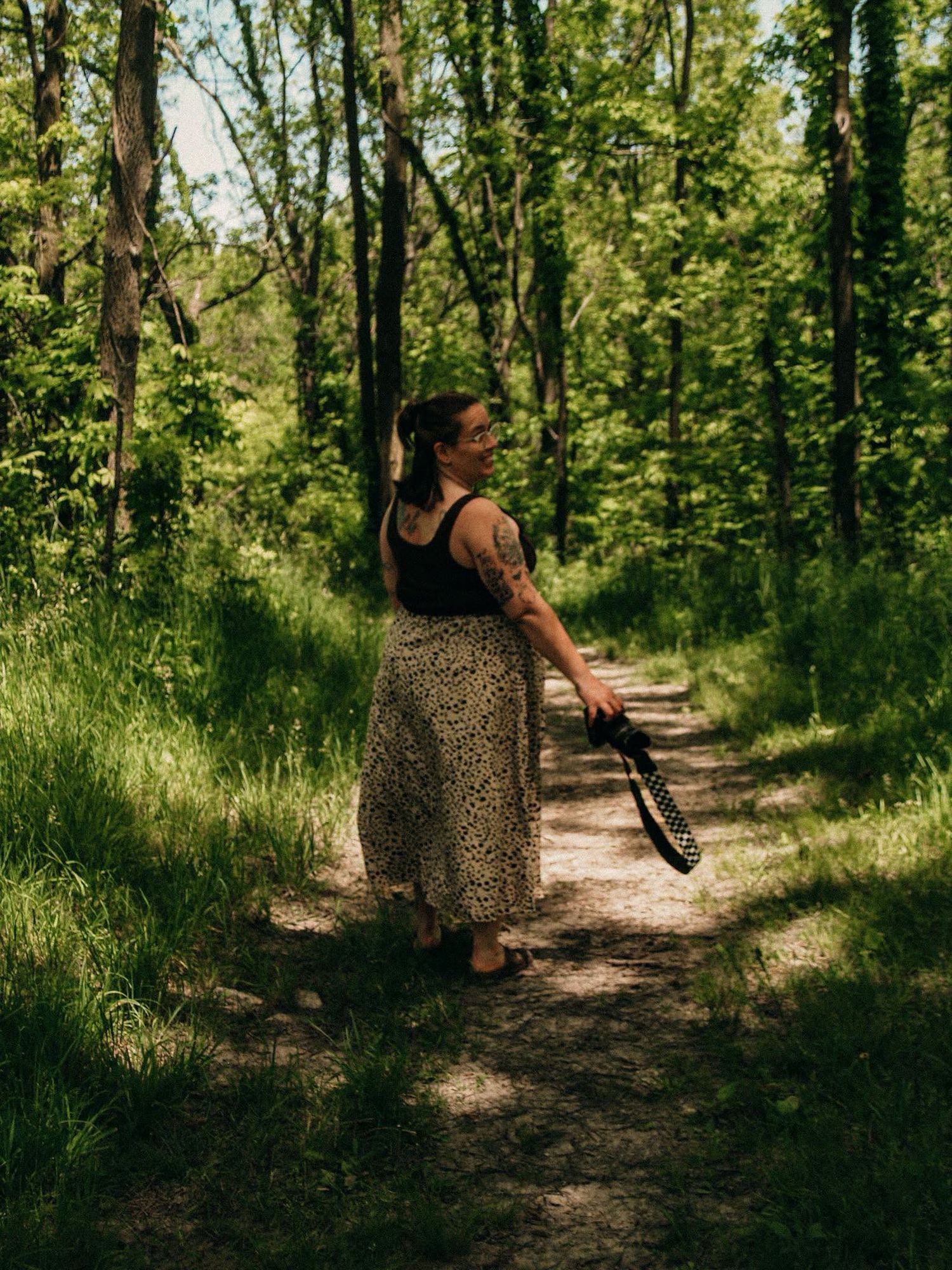 Woman in floral skirt holding guitar strap on a sunlit forest trail surrounded by lush green trees.
