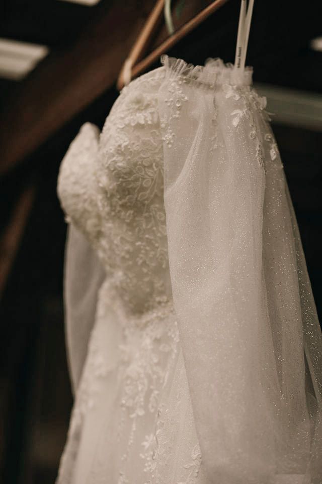 White lace wedding dress with floral appliques and tulle veil hanging on a wooden hanger in soft light.