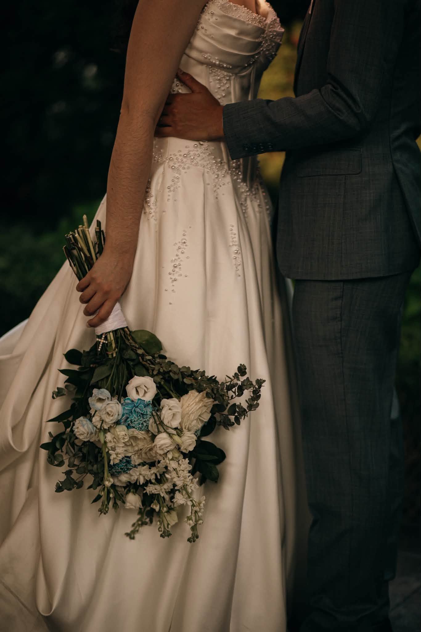 Bride in white gown holding blue and white floral bouquet with groom in gray suit embracing her waist outdoors.