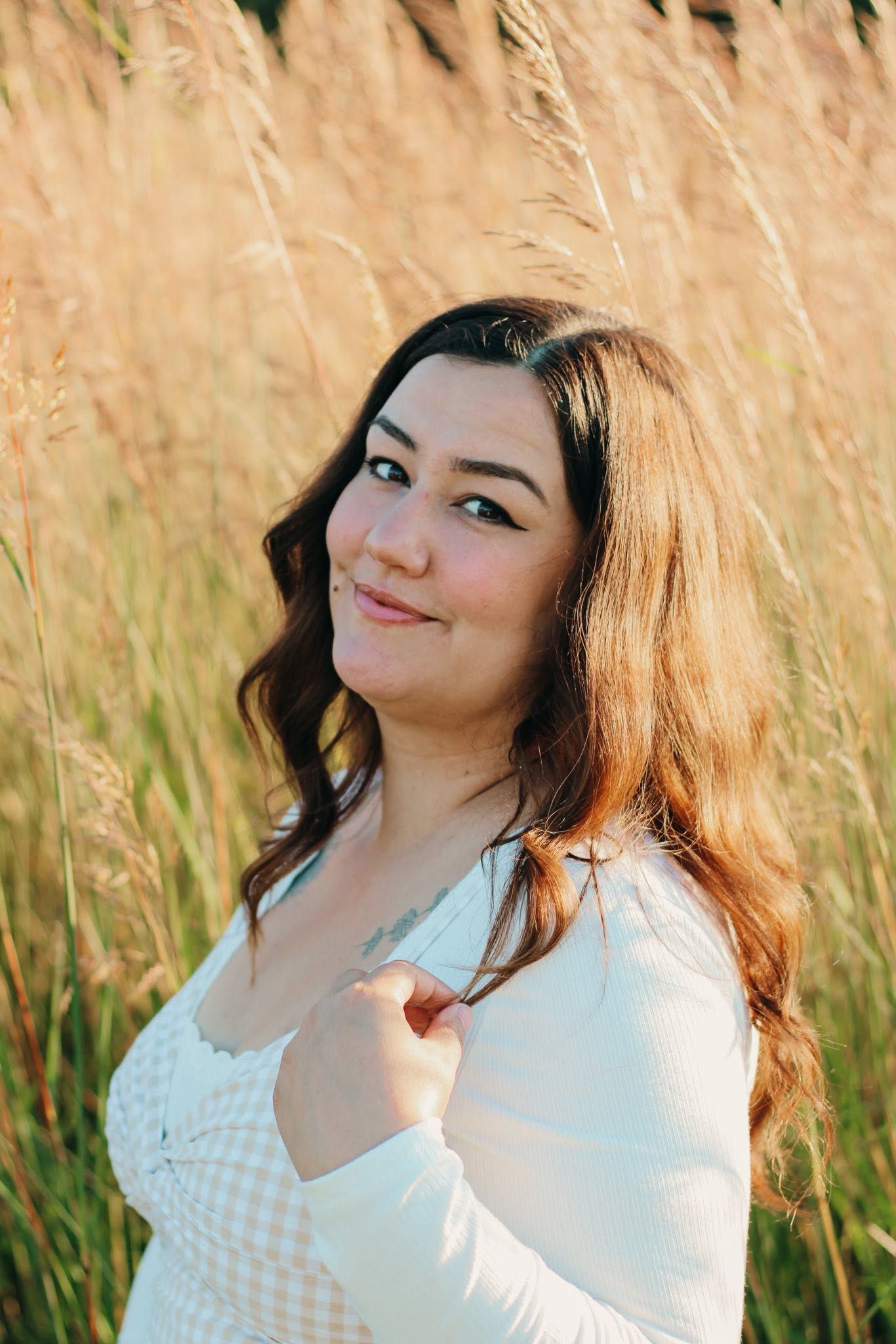 Young woman with dark wavy hair smiles softly in golden sunlit tall grass field.