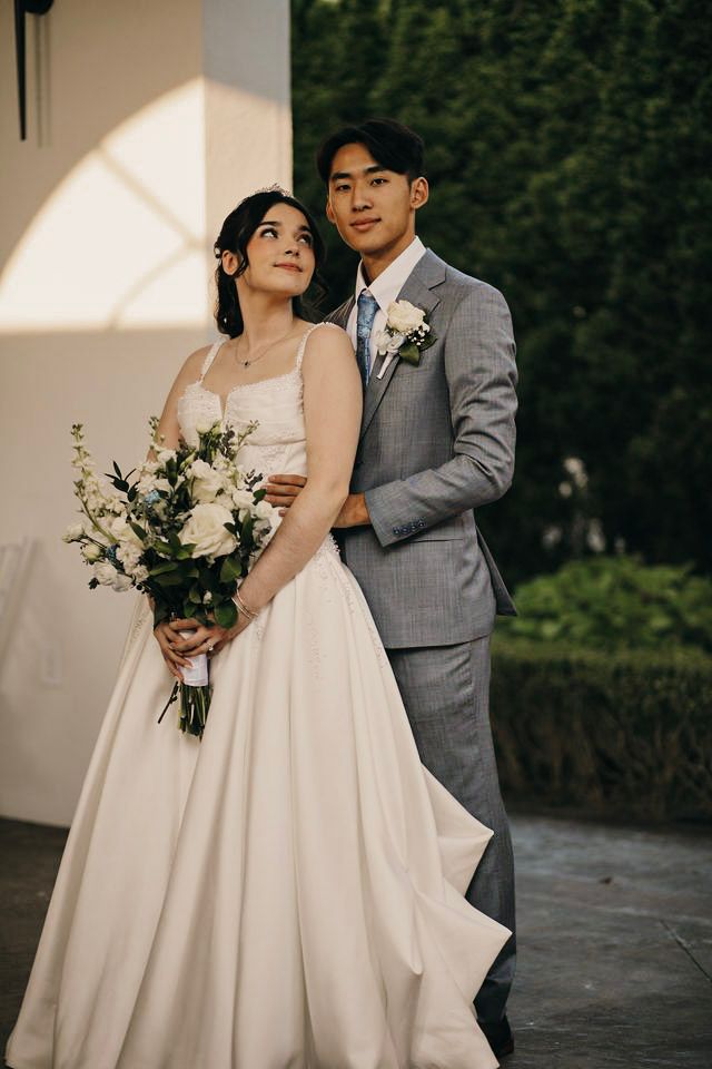 A bride in a white gown holding a floral bouquet poses with a groom in a gray suit outdoors.