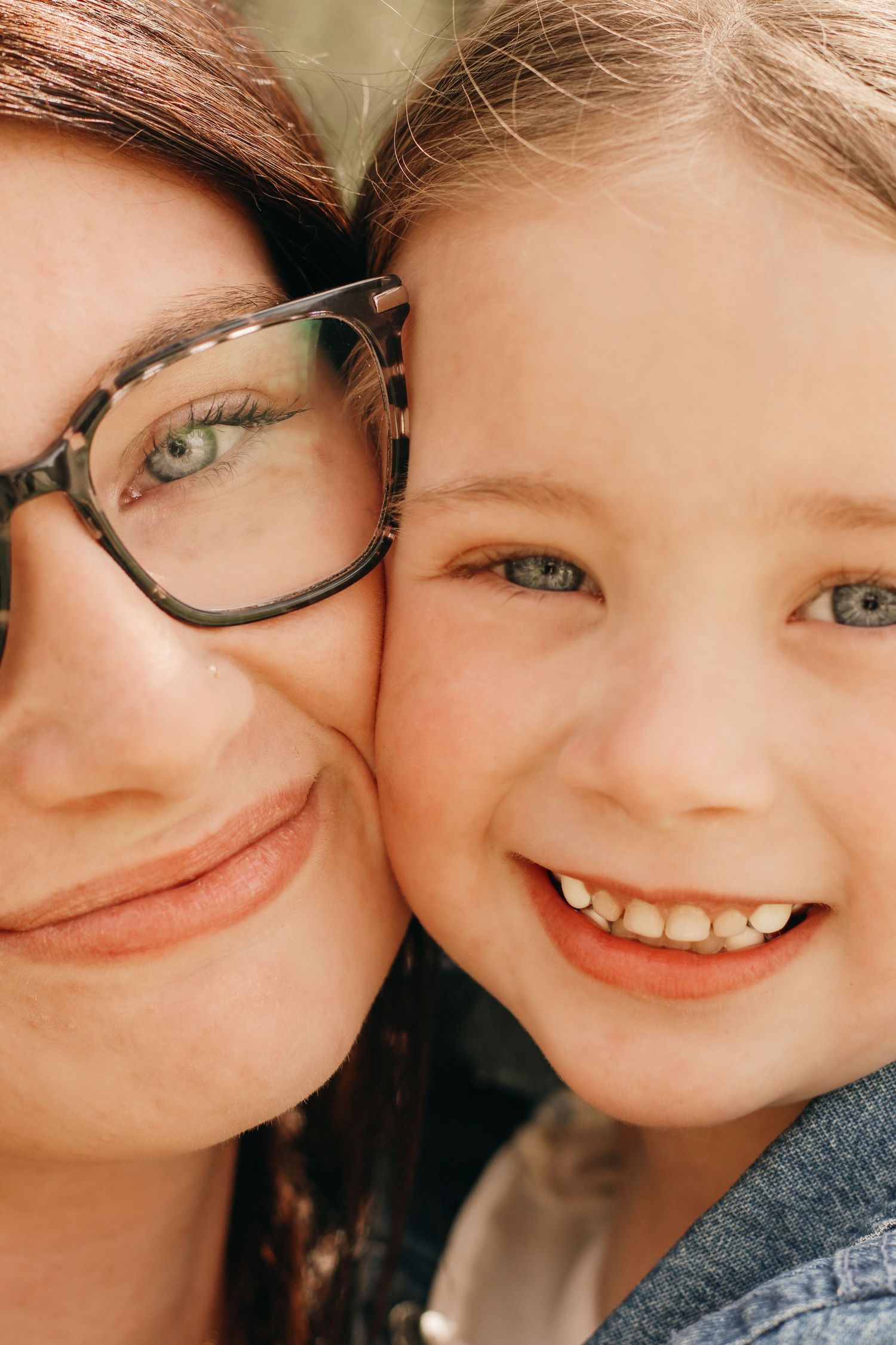 Close-up portrait of smiling mother with glasses and young daughter with blue eyes cheek to cheek.