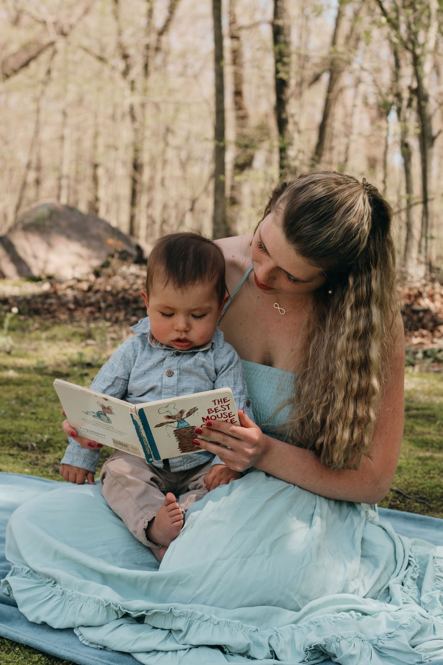 A young mother reading a picture book to her infant baby outdoors on a blue blanket.