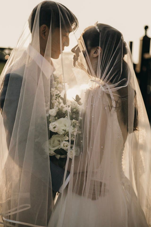 A bride and groom share an intimate moment under a flowing white veil, bride holding a white floral bouquet.