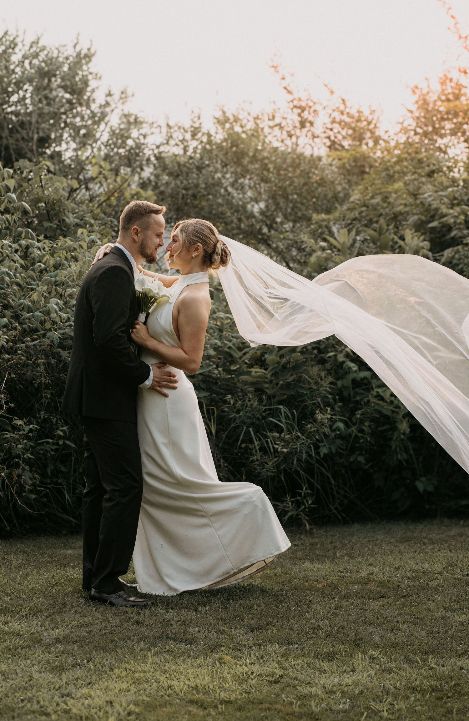 Bride and groom embrace in garden at golden hour, veil flowing in wind, lush green trees in background.