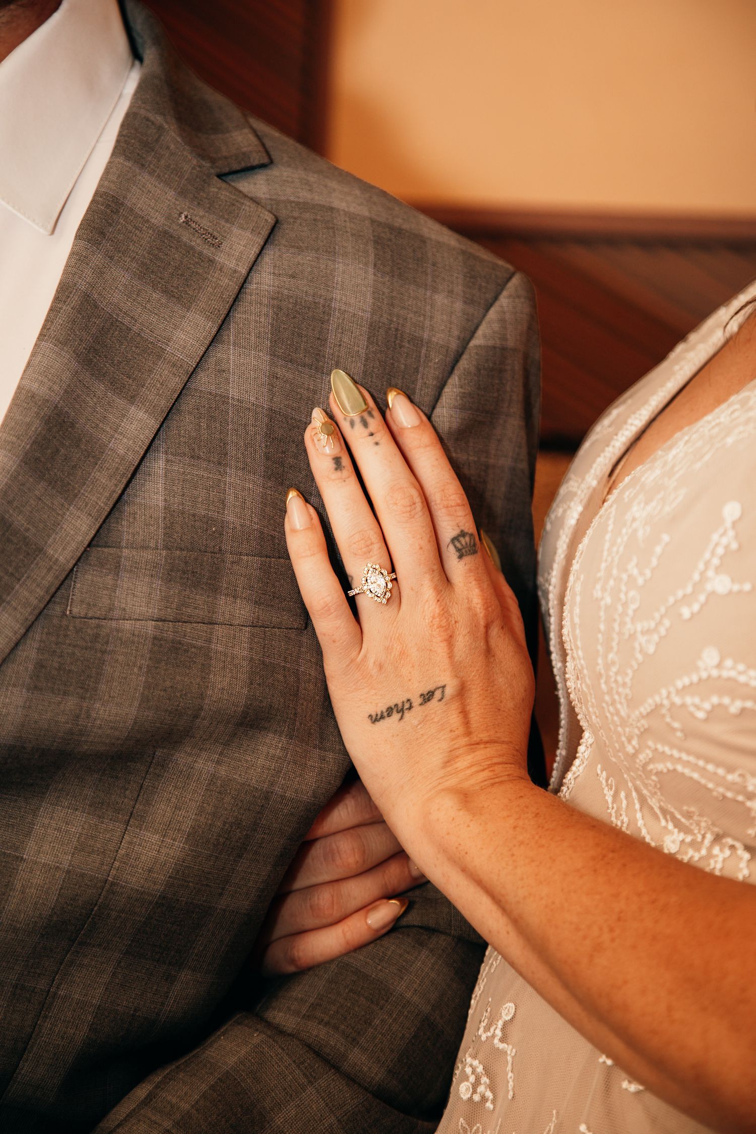 Close-up of bride's tattooed hand with oval engagement ring resting on groom's gray plaid suit jacket.