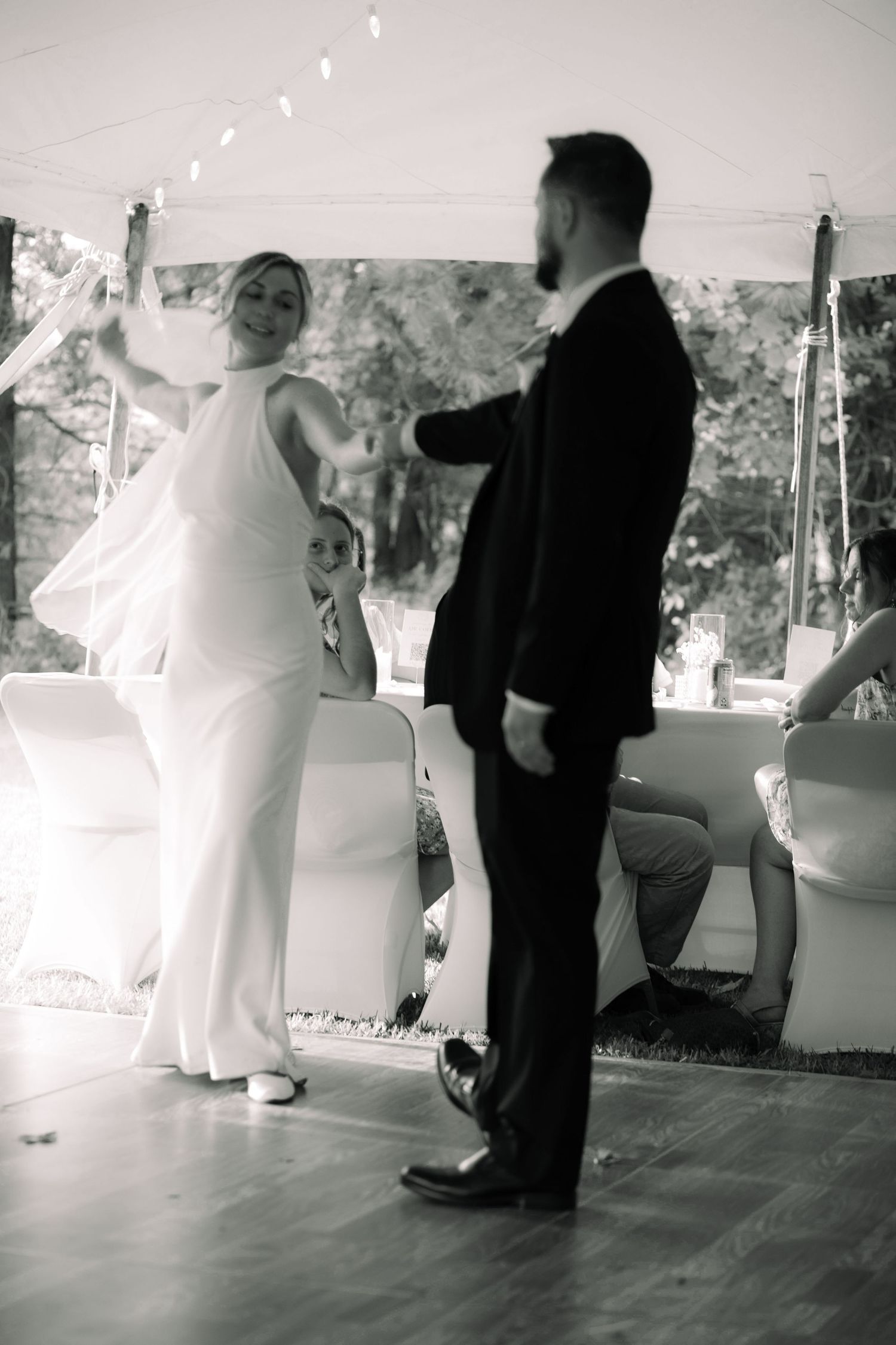 Black and white photo of bride and groom sharing first dance under a tent with string lights.