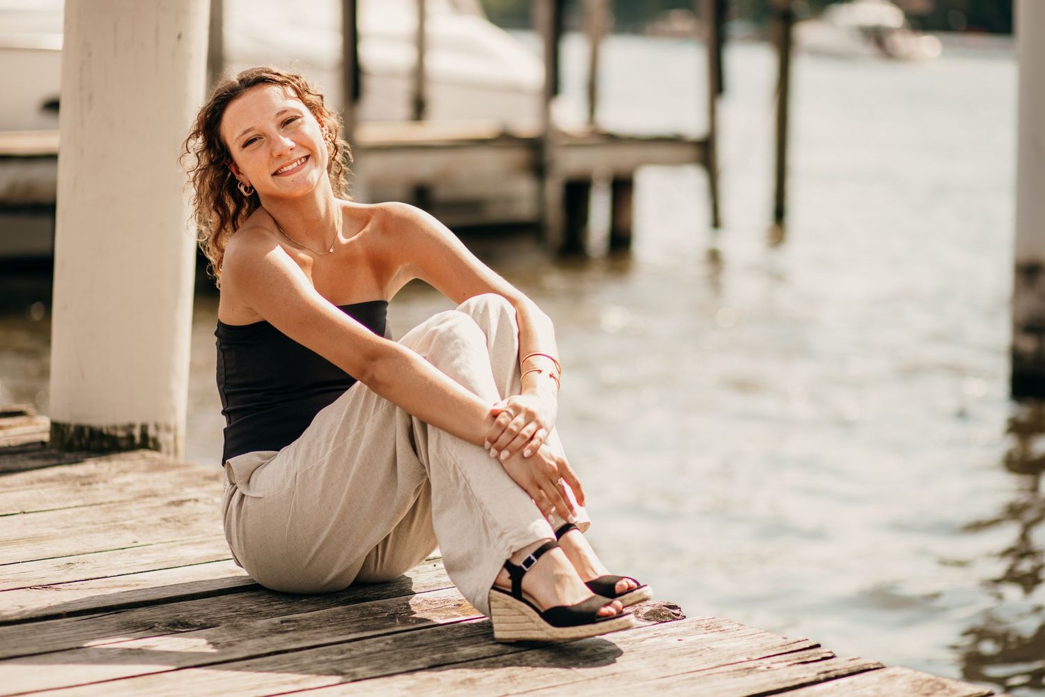 Smiling woman with curly hair sitting on a wooden dock by the water in summer.