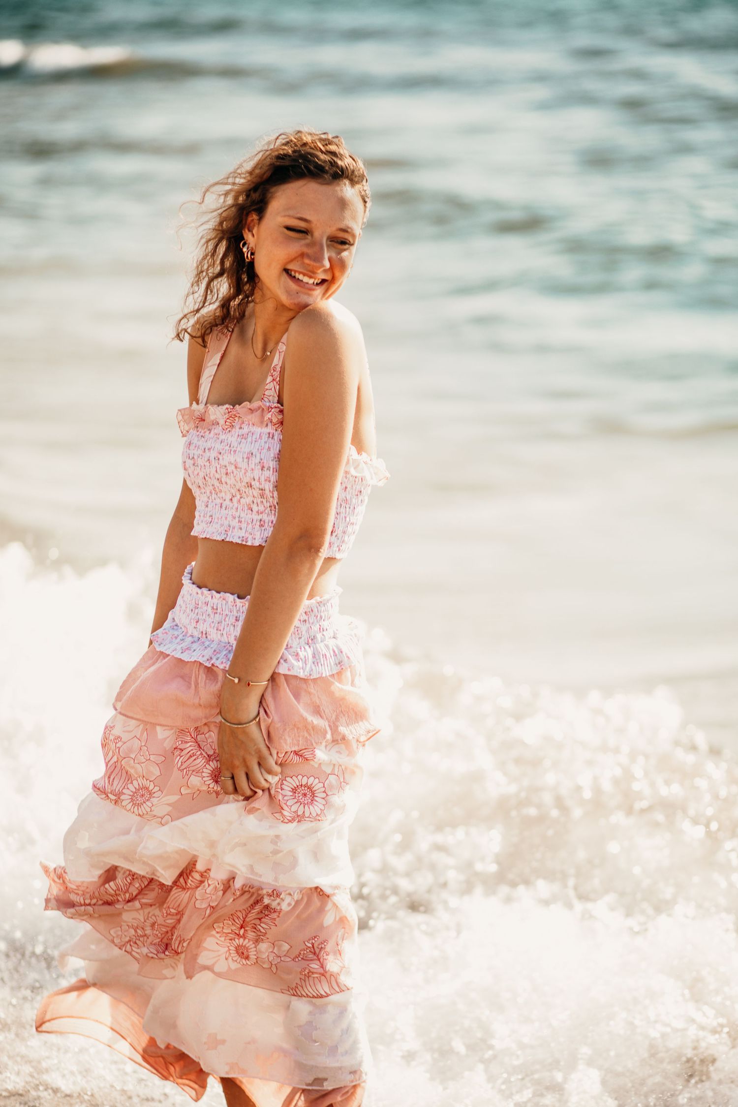 Smiling woman in pink ruffled crop top and maxi skirt standing in ocean waves on sunny beach.