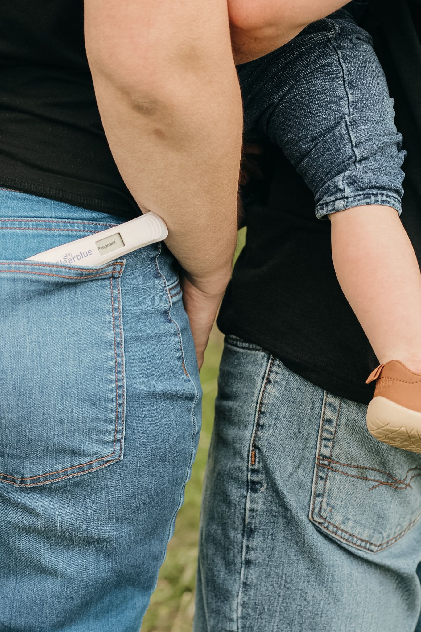 Couple in jeans holding a positive pregnancy test in back pocket outdoors.