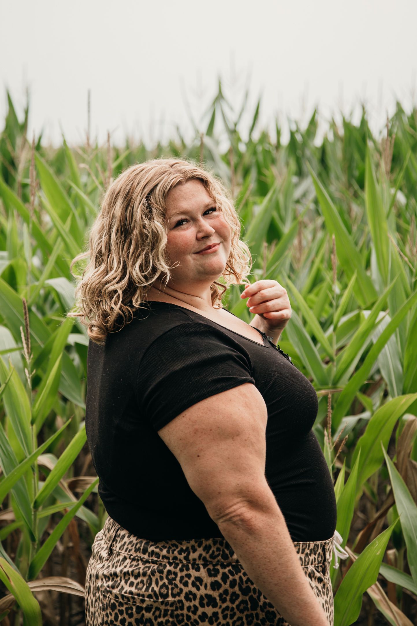 Plus-size woman with blonde curly hair posing in a green cornfield wearing a black top and leopard print skirt.