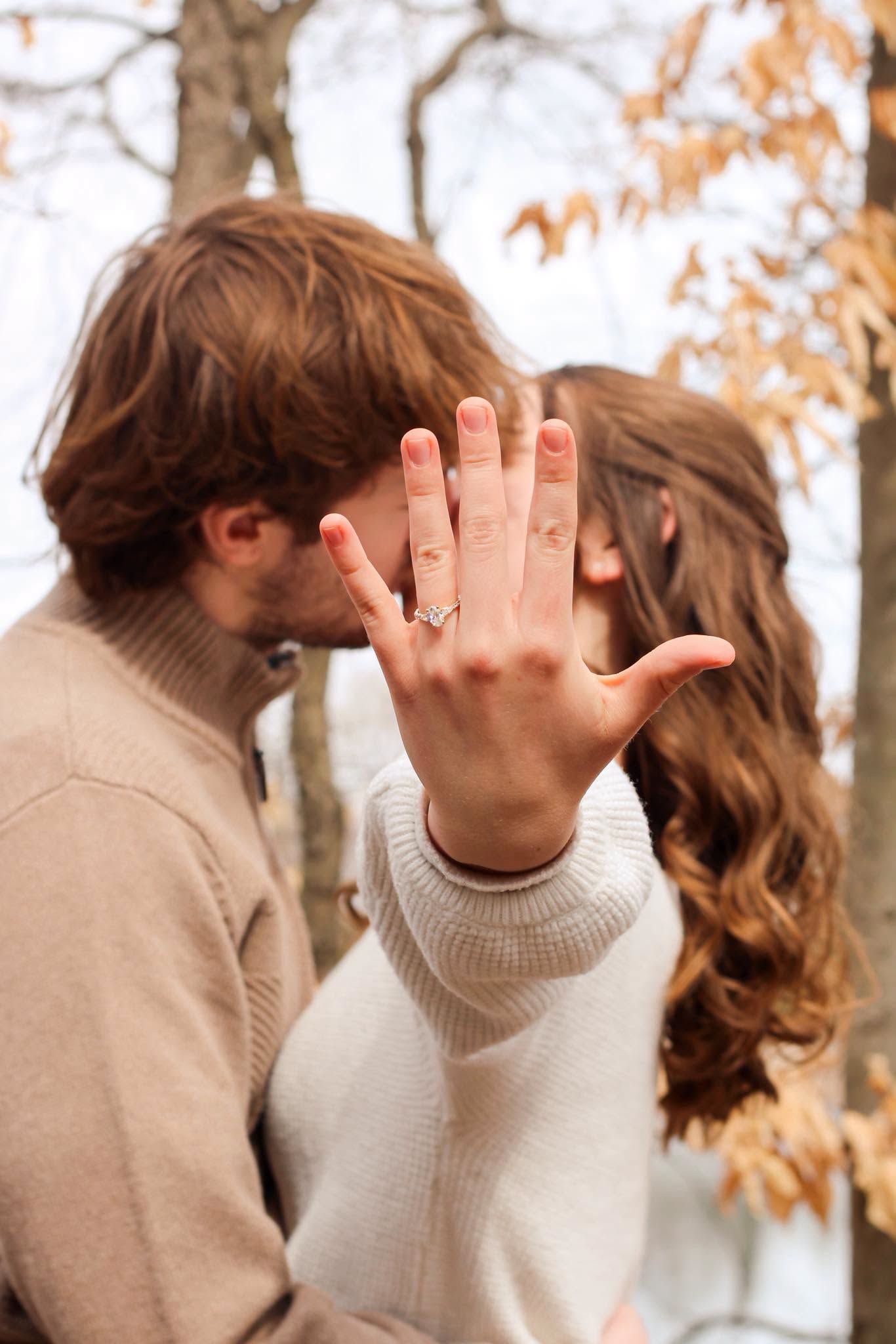 Couple kissing in autumn park while woman holds up hand displaying sparkling diamond engagement ring.