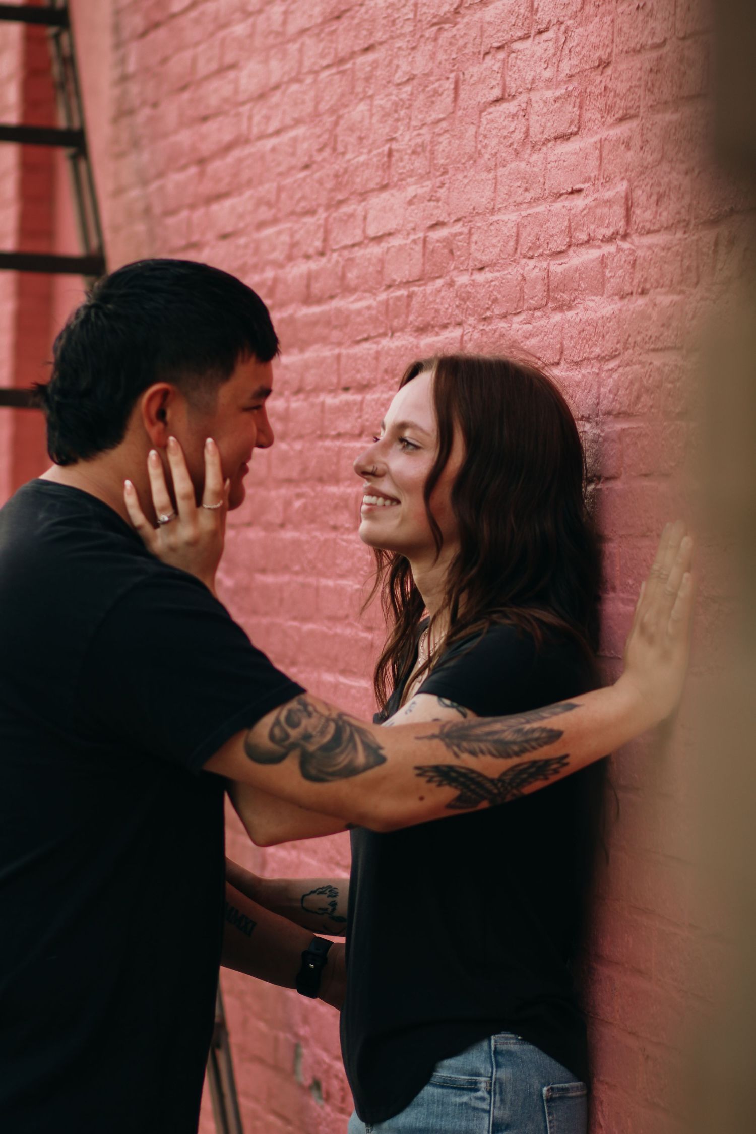 Tattooed couple sharing a romantic moment against a pink brick wall in an urban alley setting.