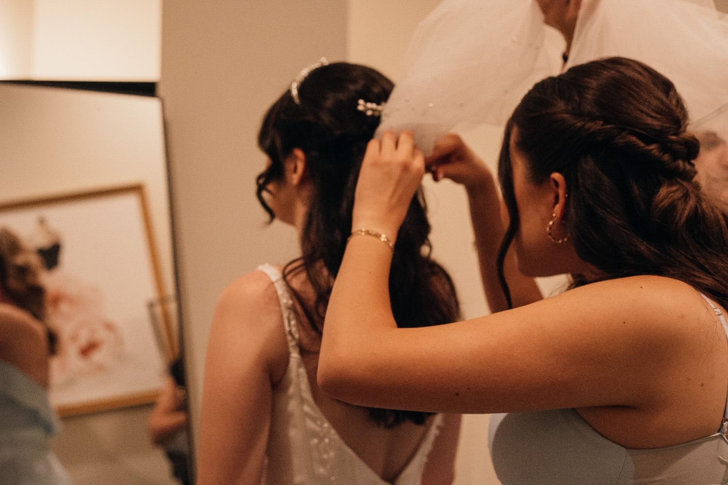 Bridesmaids helping bride with hair accessories and dress buttons in a warmly lit dressing room before the wedding ceremony.