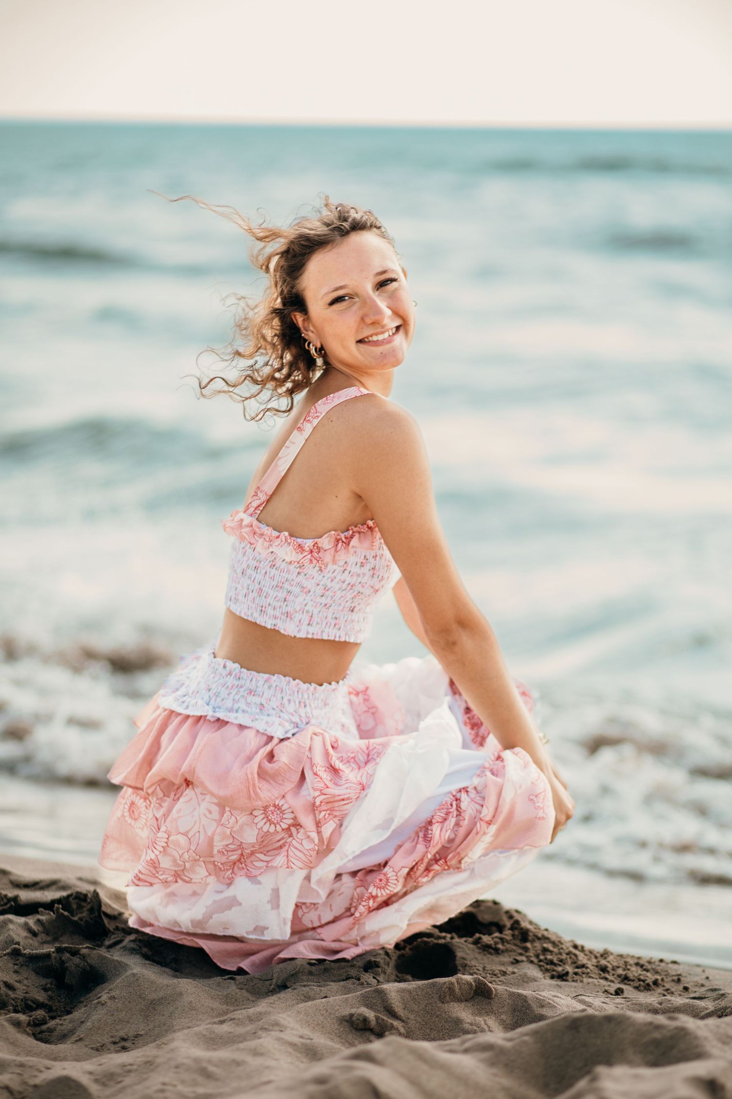 Happy woman in pink crop top and skirt sitting on sandy beach with ocean behind her.