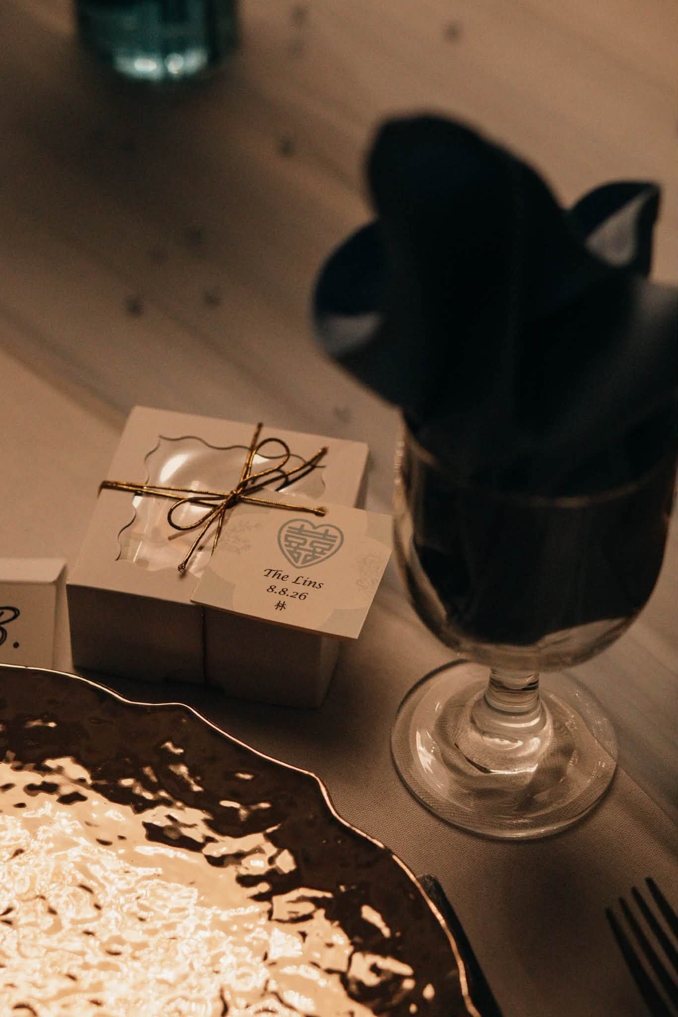 Elegant wedding favor box with twine bow beside a wine glass and gold decorative plate on wooden table.