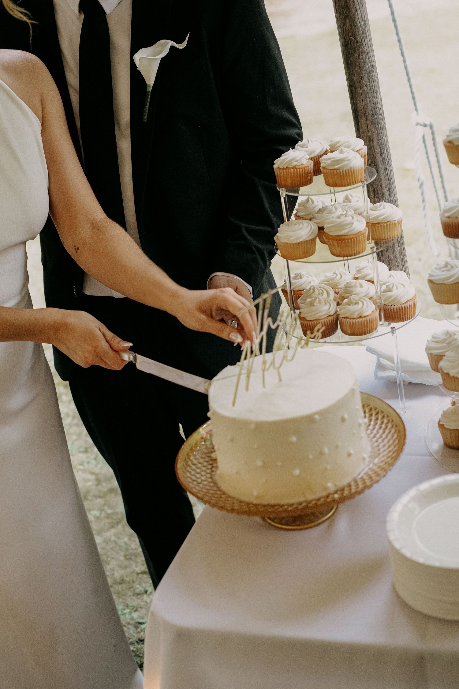 Bride and groom cutting a white wedding cake beside a cupcake tower display at their wedding reception.