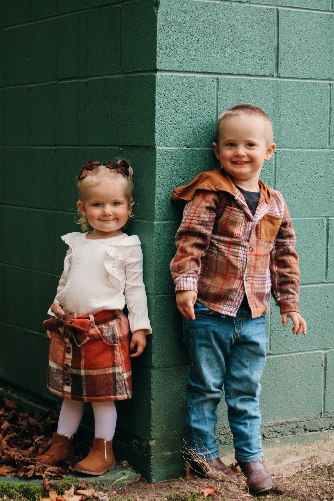 Two toddlers in matching plaid outfits smile against a green brick wall in an adorable fall photo session.