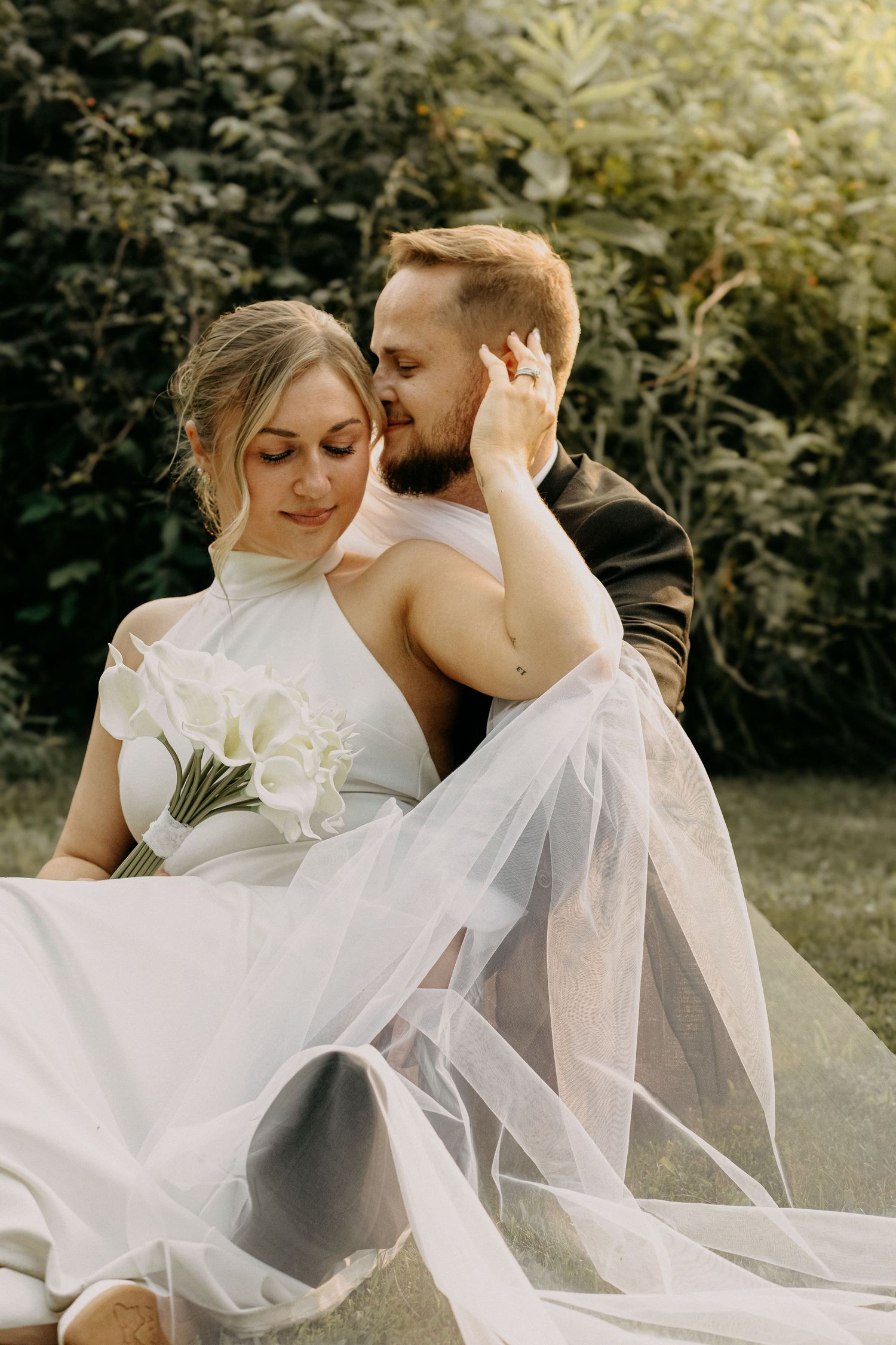 Groom kisses bride on cheek while holding her in his arms, bride holds white calla lily bouquet.