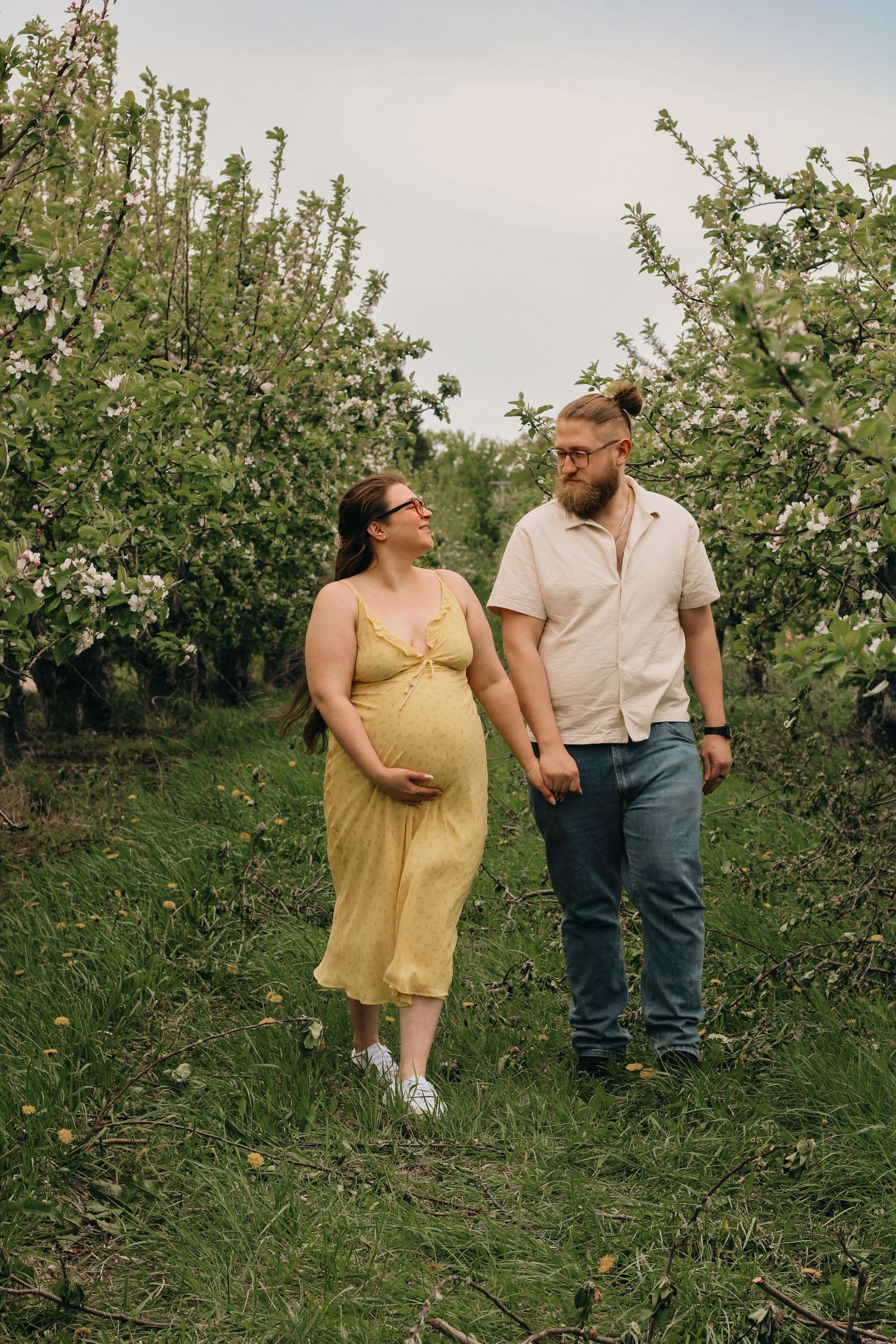 Pregnant woman in yellow dress and partner walk hand-in-hand through blossoming apple orchard.