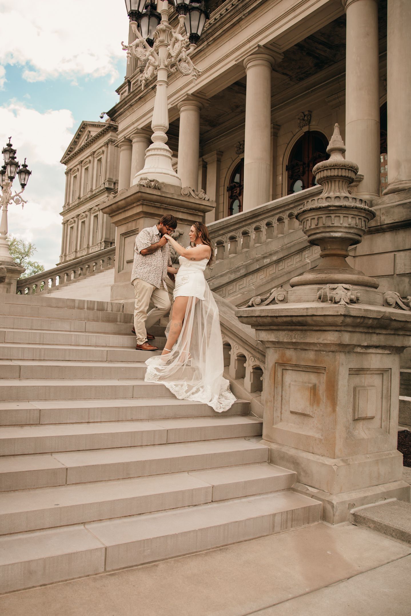 Bride and groom pose on grand stone steps of a classical building with columns on their wedding day.