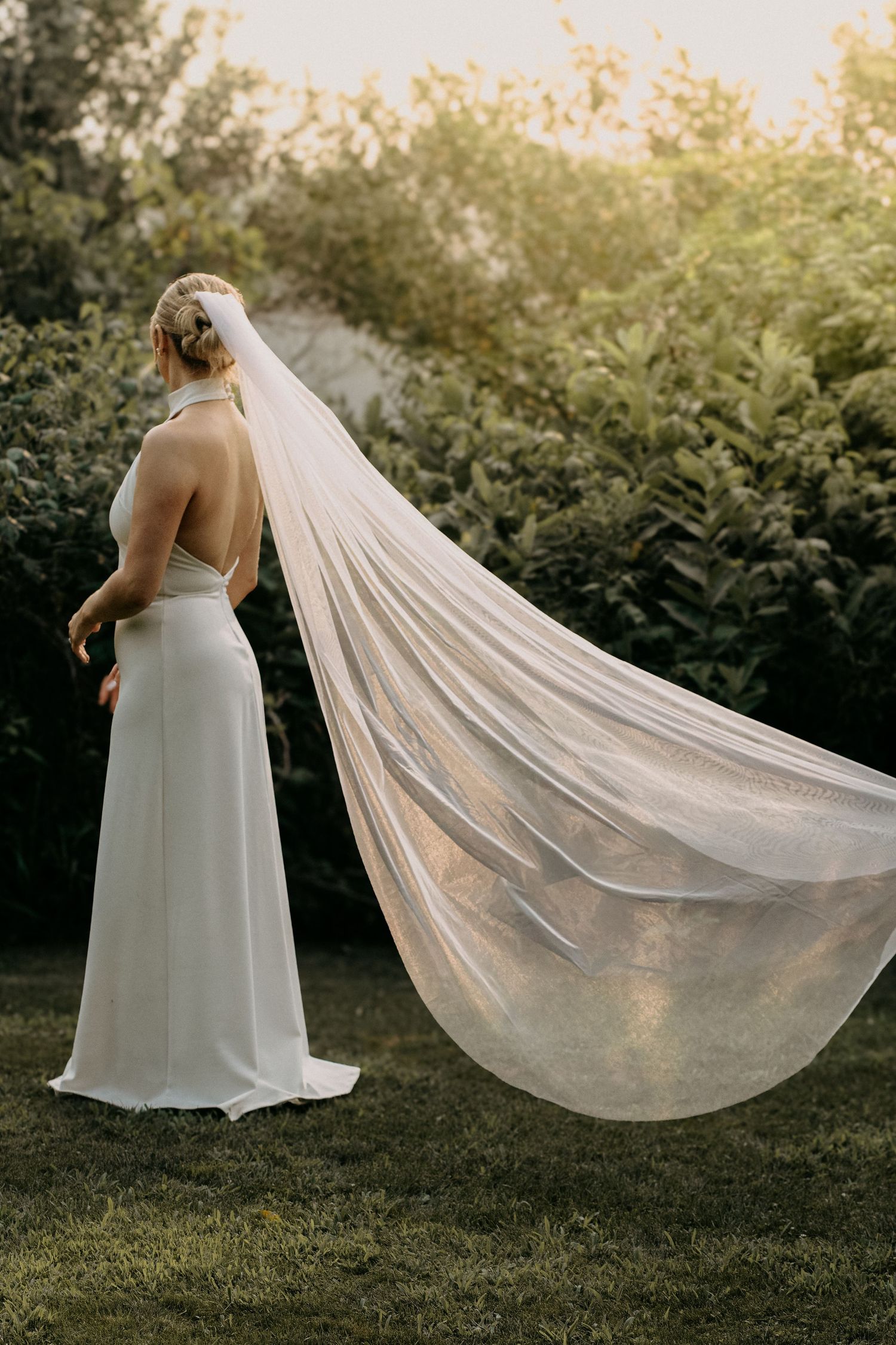 Bride in elegant white halter gown with dramatic cathedral veil flowing in garden sunset.
