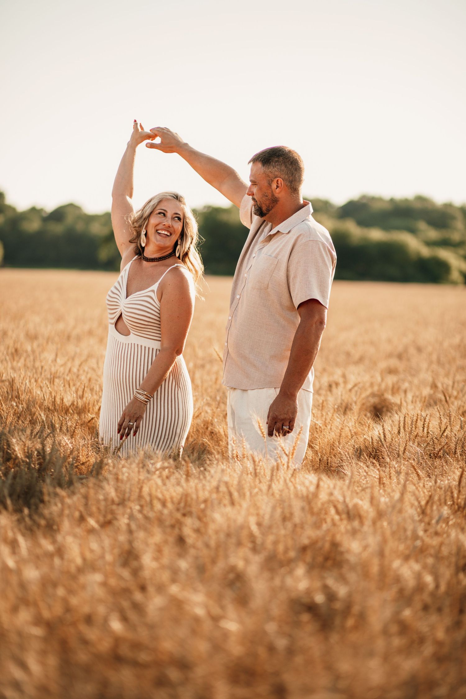 Couple dancing joyfully in a golden wheat field at sunset, woman in striped dress twirling.