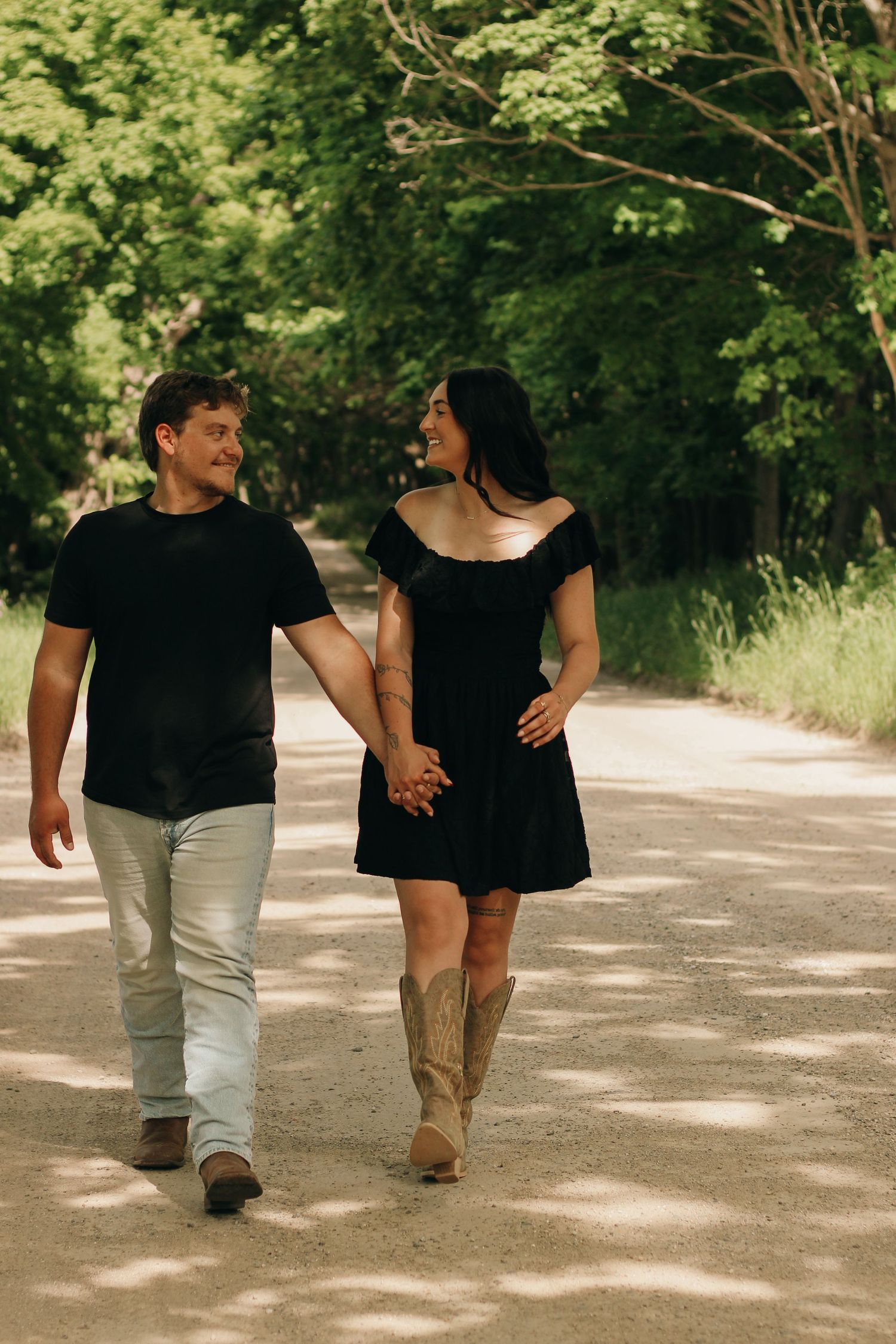 Happy couple holding hands walking down a tree-lined path, woman wearing black dress and cowboy boots.