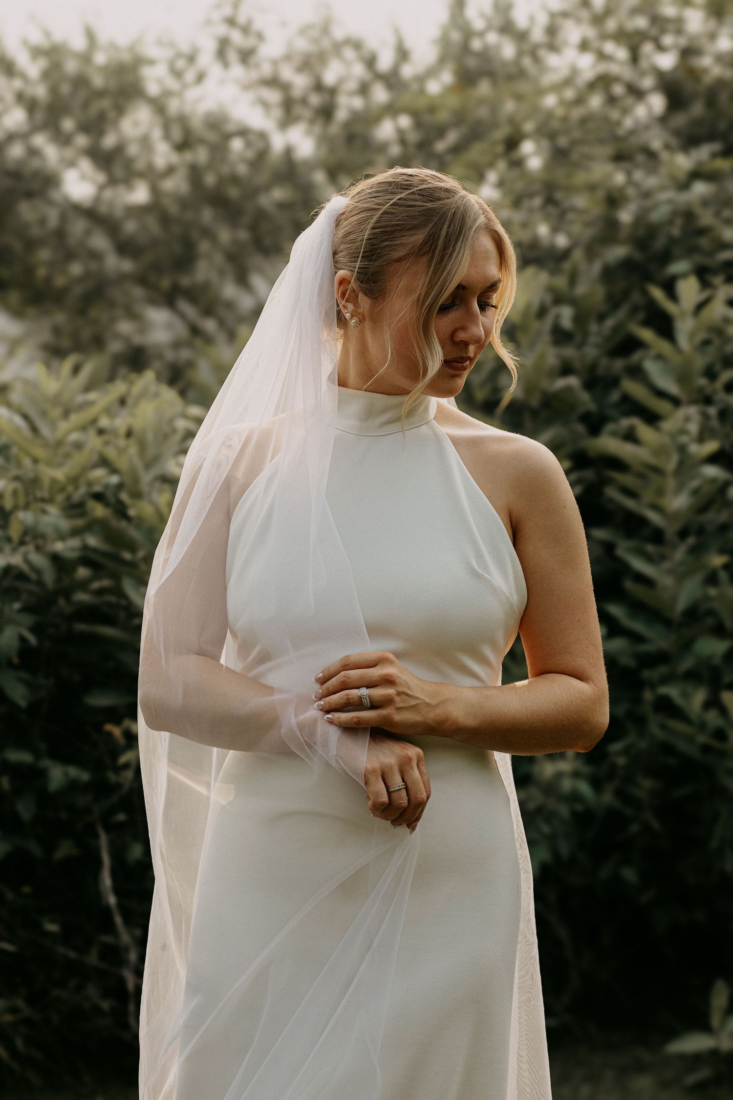 Elegant bride in sleek white halter gown and veil gazes down in lush garden.