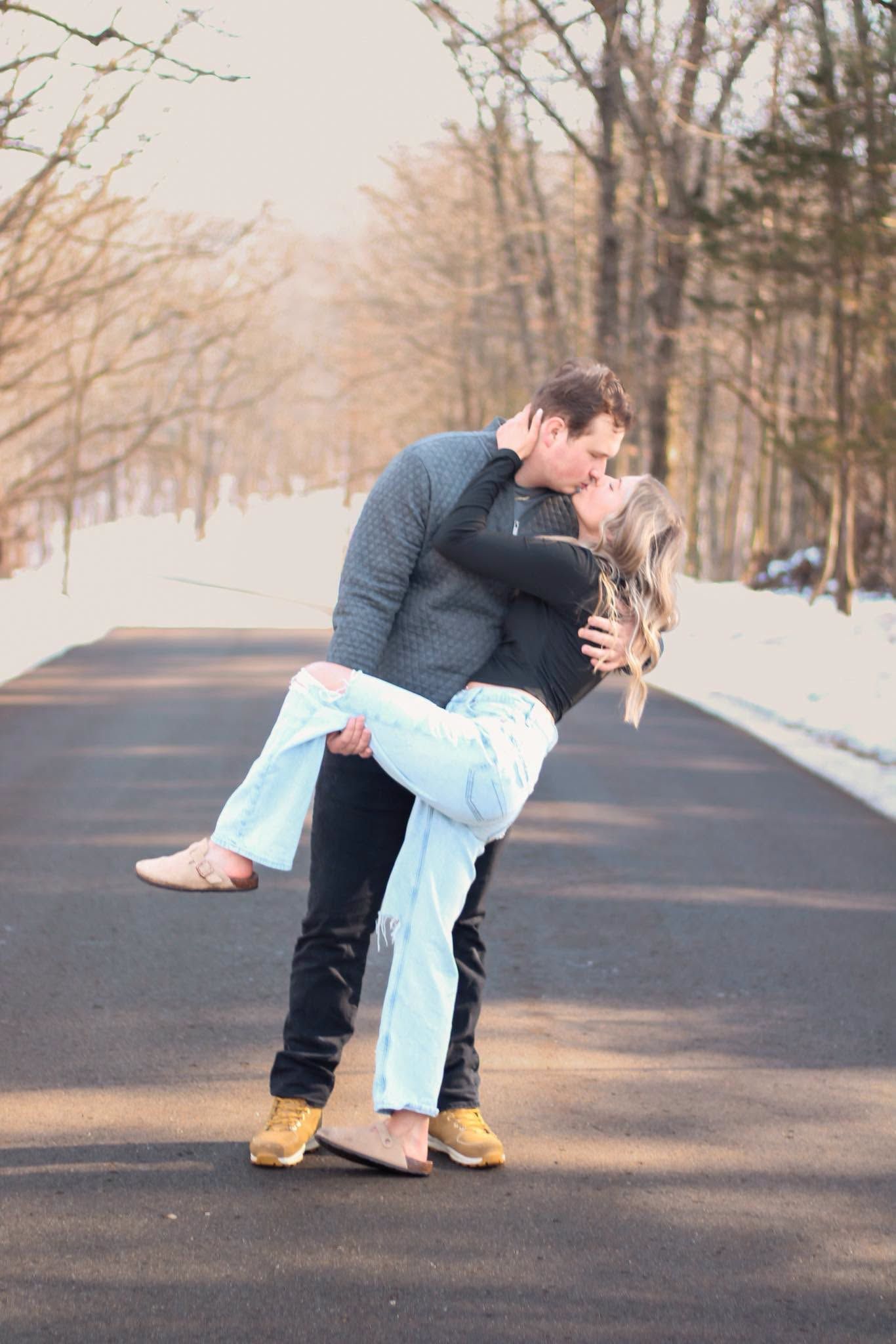 Couple sharing a romantic kiss on a snowy winter road, man lifting woman in light blue jeans among bare trees.