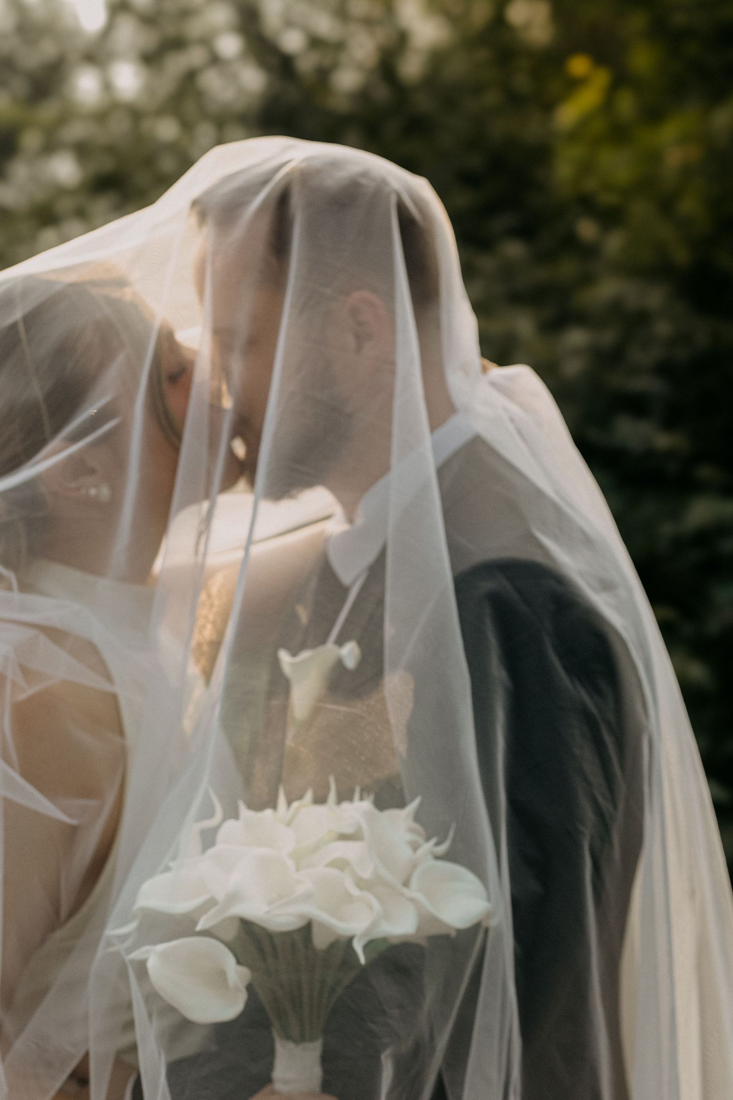 Bride and groom sharing a kiss under a flowing white veil holding white calla lily bouquet outdoors.