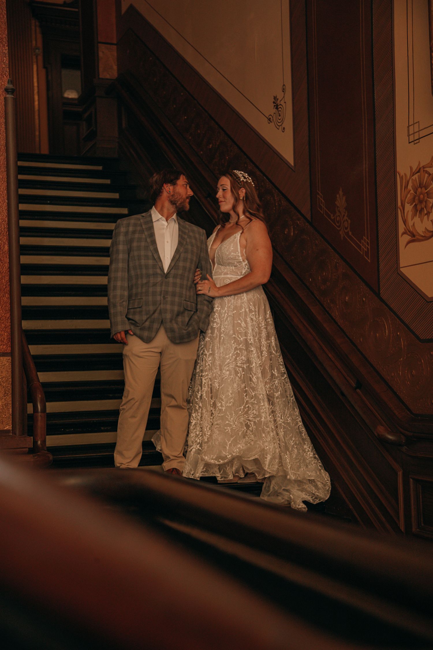 A bride in a lace gown and groom in a plaid suit share a romantic moment on a grand staircase.