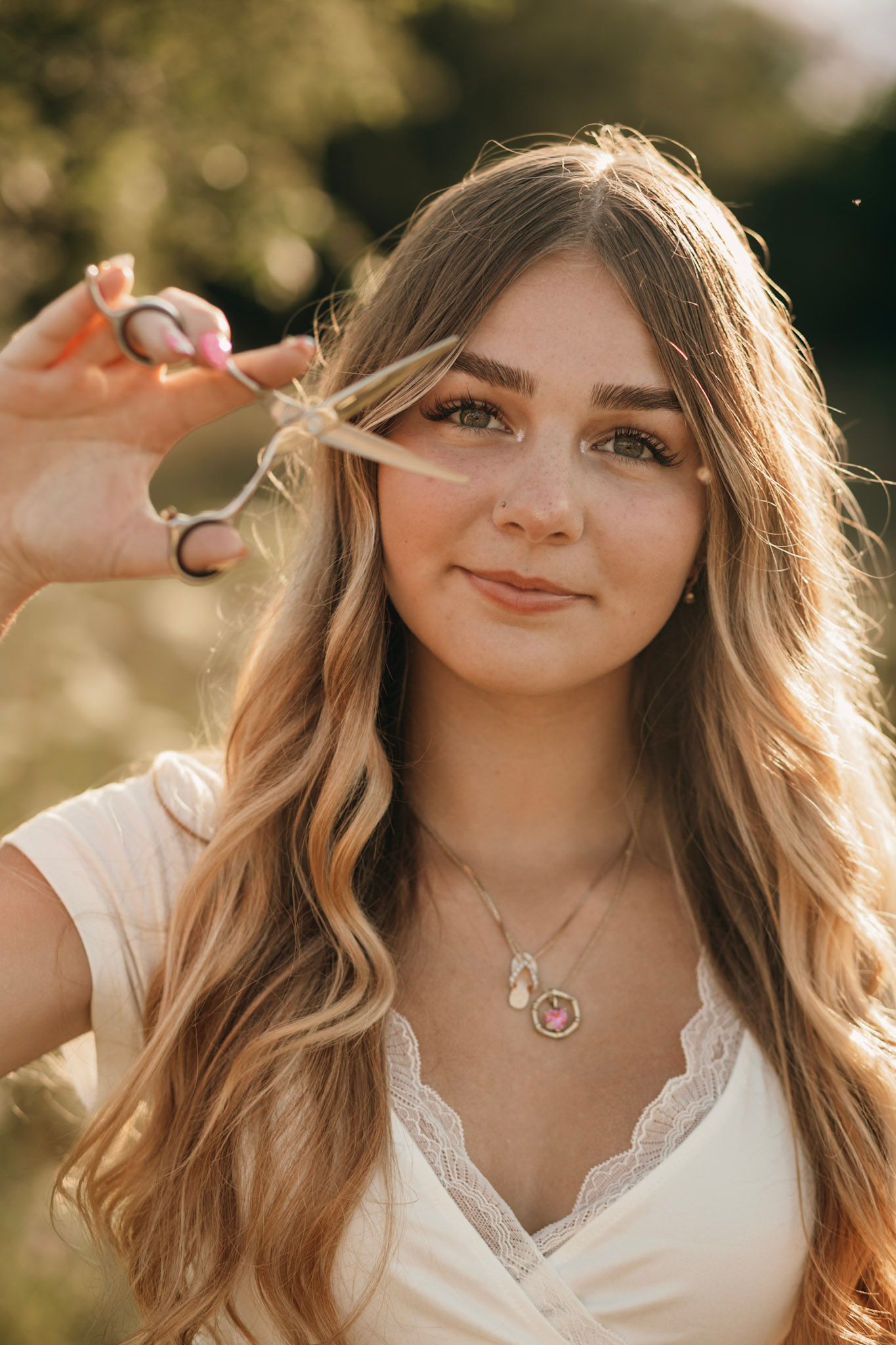 Young woman with long wavy hair holds scissors near her face, wearing a white top and pink pendant necklace outdoors.