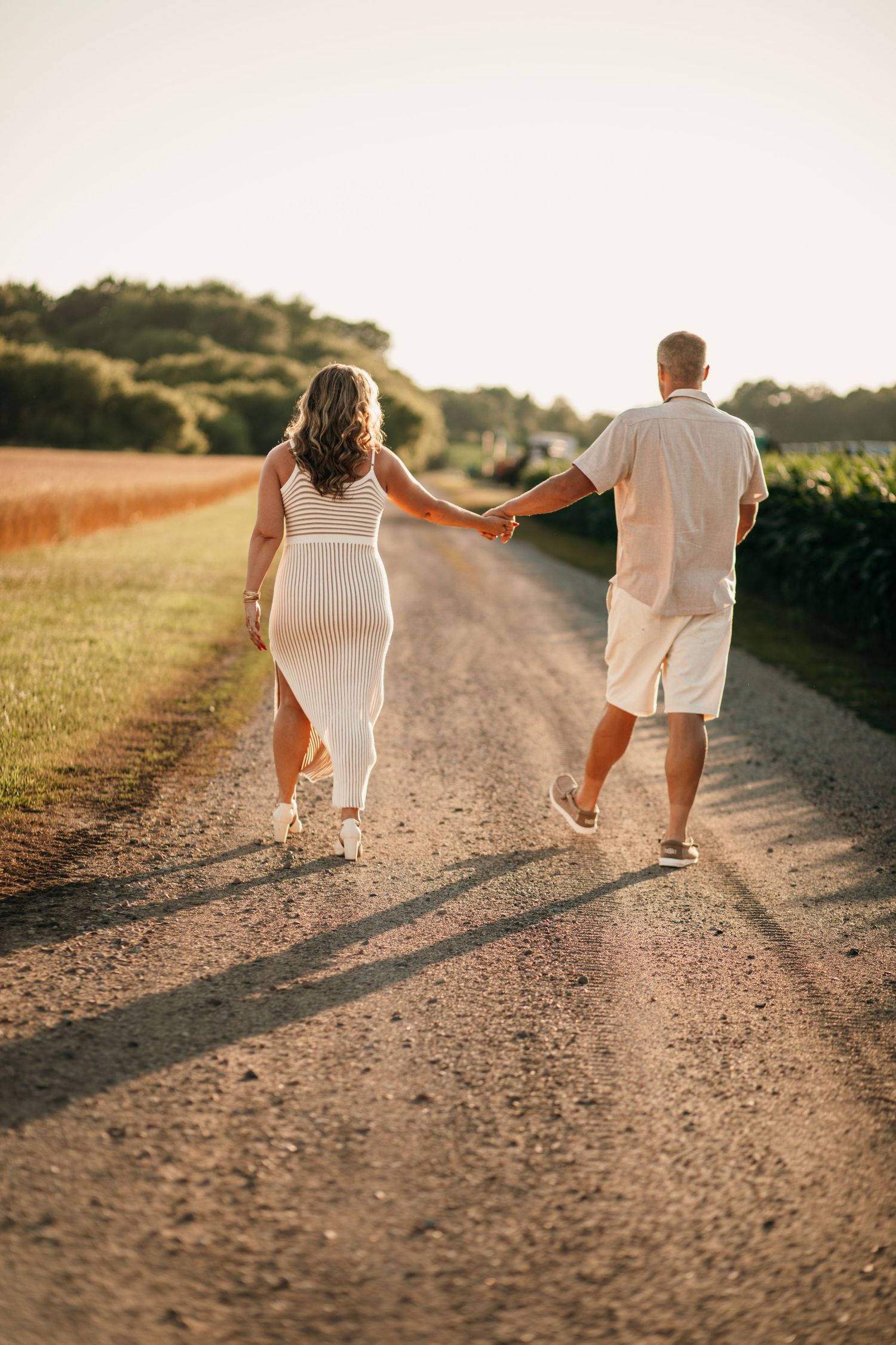 Couple in white outfits holding hands walking down a rural dirt road at golden hour sunset.